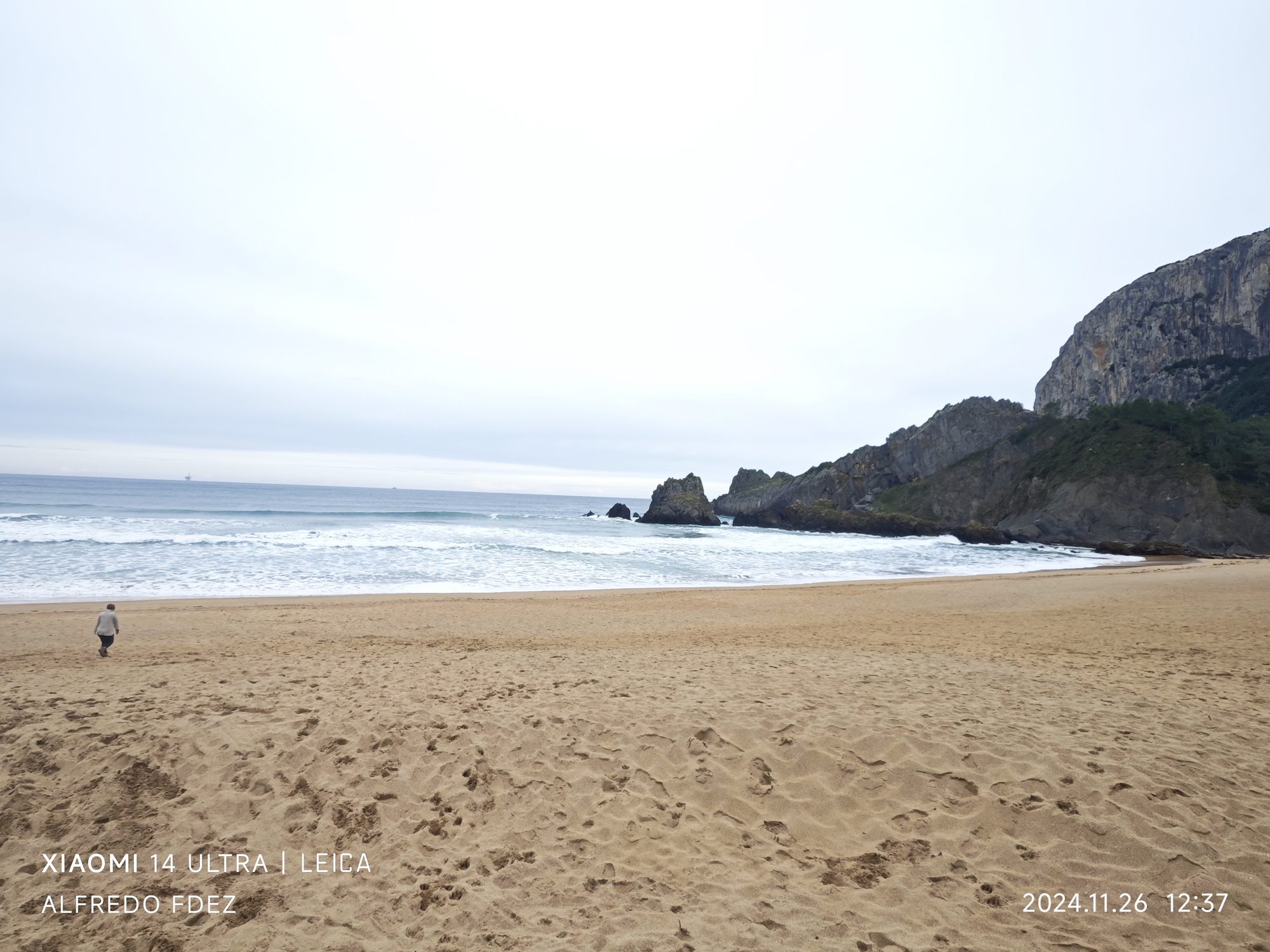 Playa de arena dorada con olas del mar y acantilados rocosos bajo cielo nublado.