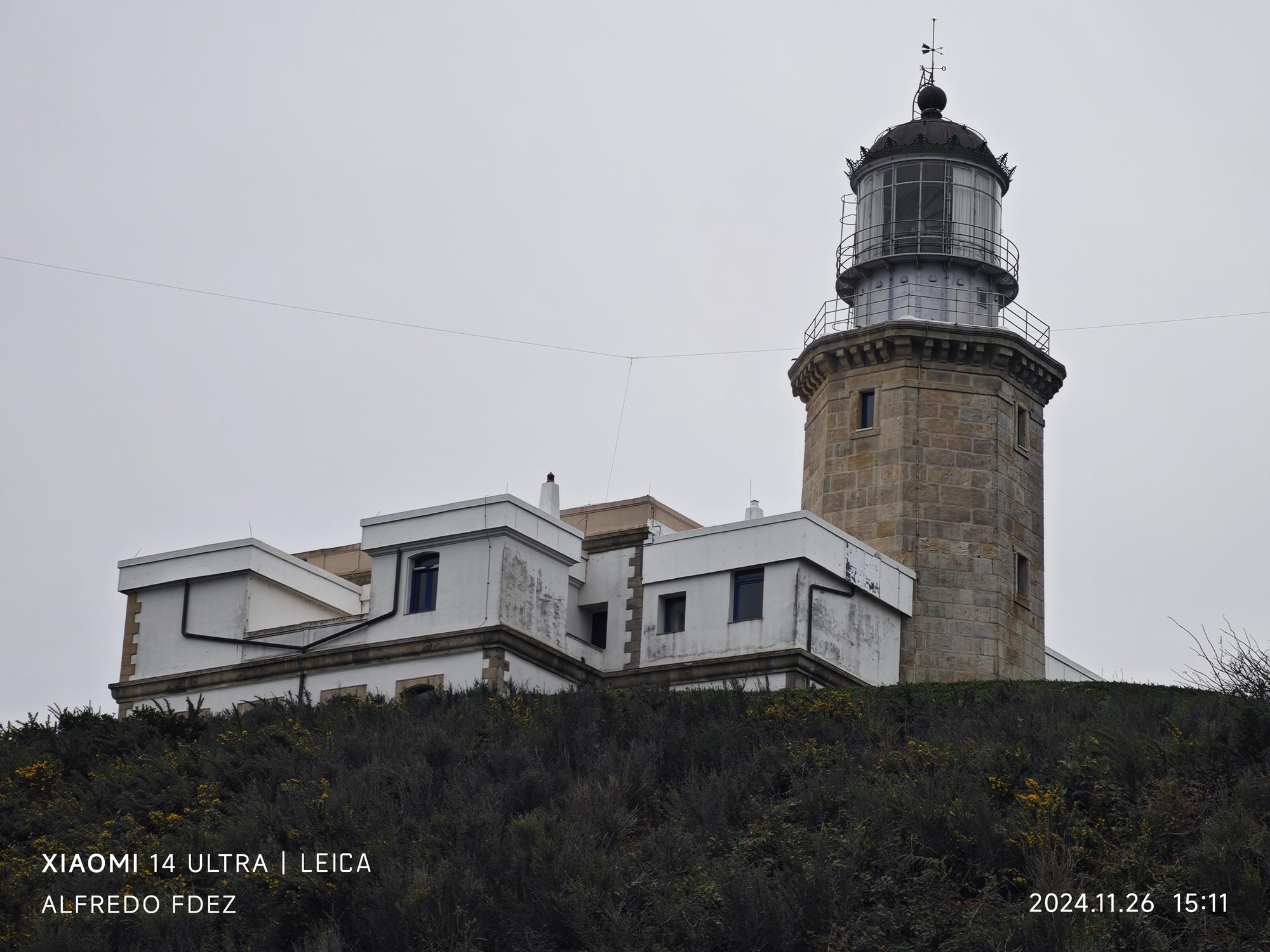 Faro de piedra antiguo con edificios blancos sobre una colina en un día nublado.