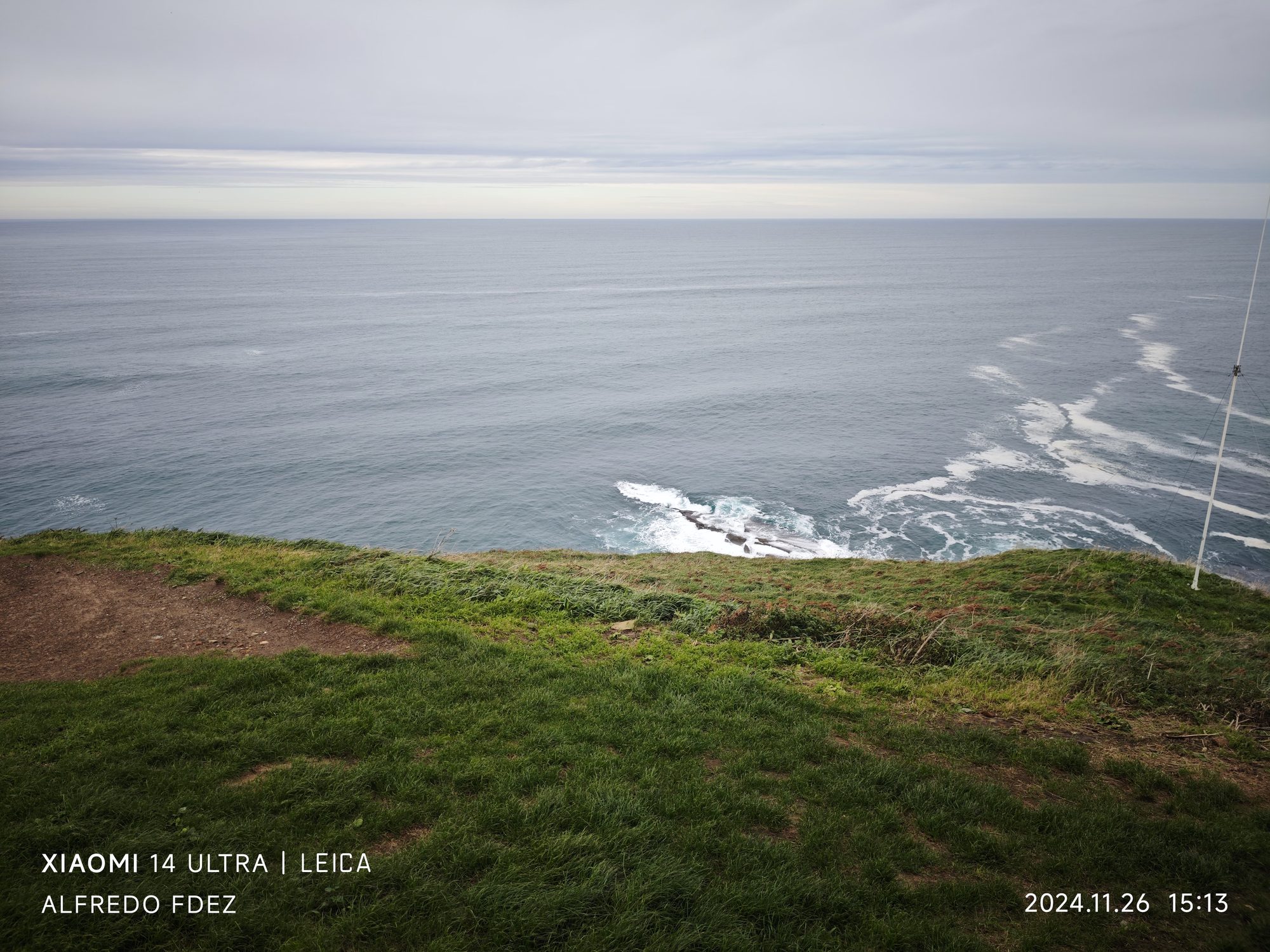 Acantilado de hierba verde frente al océano Cantábrico bajo un cielo gris nublado.