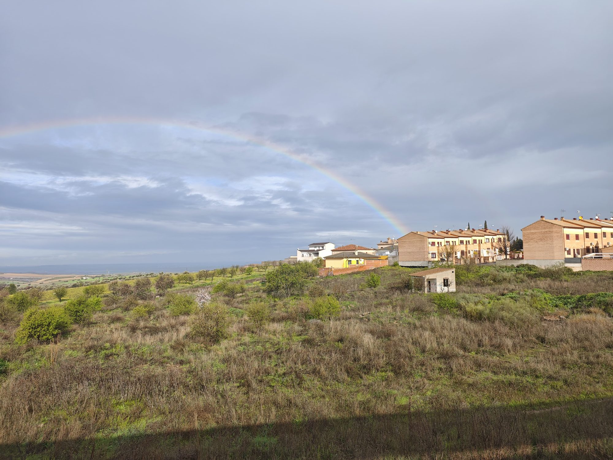 Arcoíris brillante cruzando el cielo nublado sobre el paisaje rural y casas de Bargas.