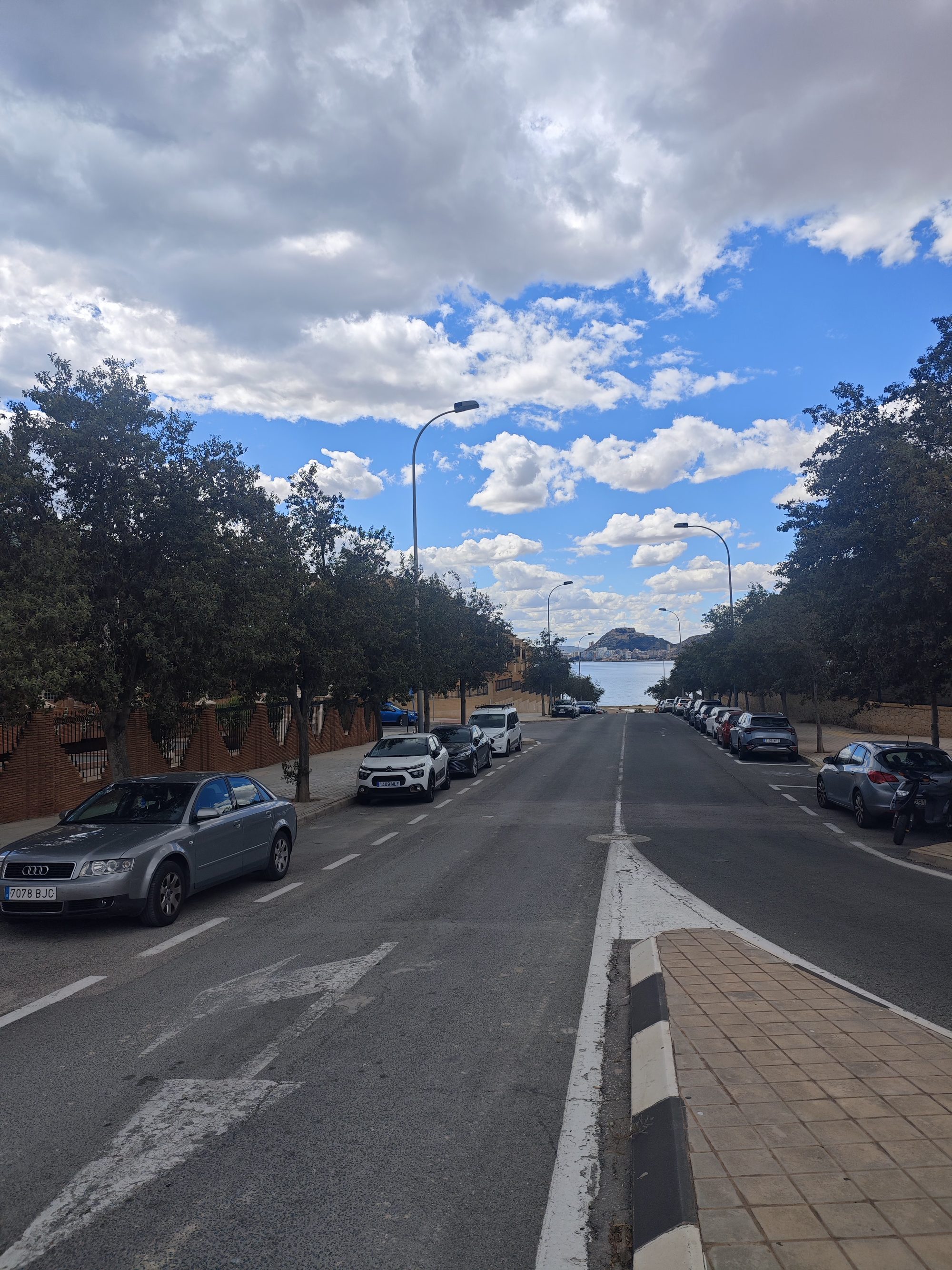 Calle asfaltada en pendiente hacia el mar con coches aparcados y cielo nublado.