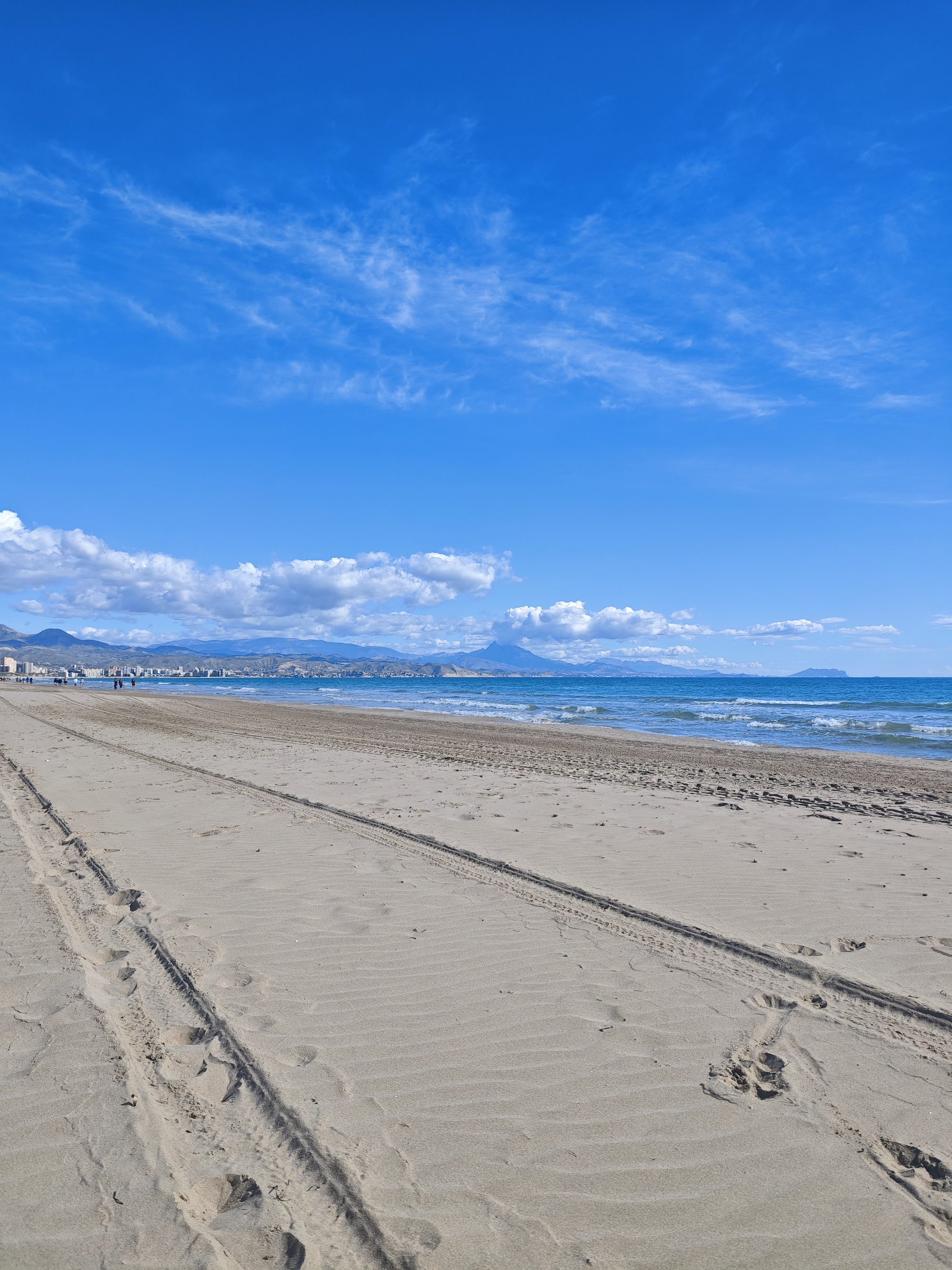 Amplia playa de arena en Alicante con huellas, mar azul y montañas en el horizonte.