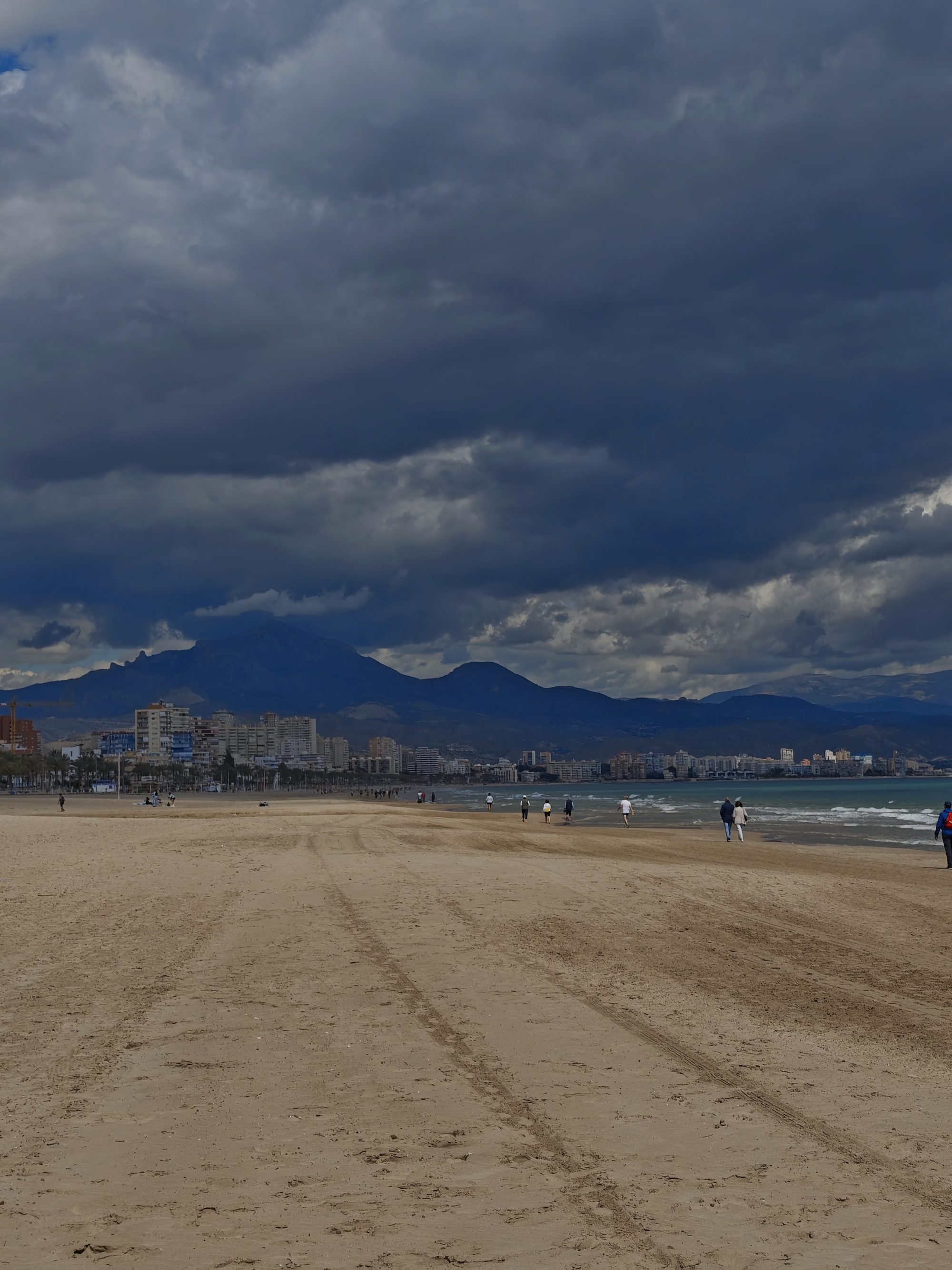 Amplia playa de Alicante con mar, edificios costeros y nubes oscuras sobre las montañas.