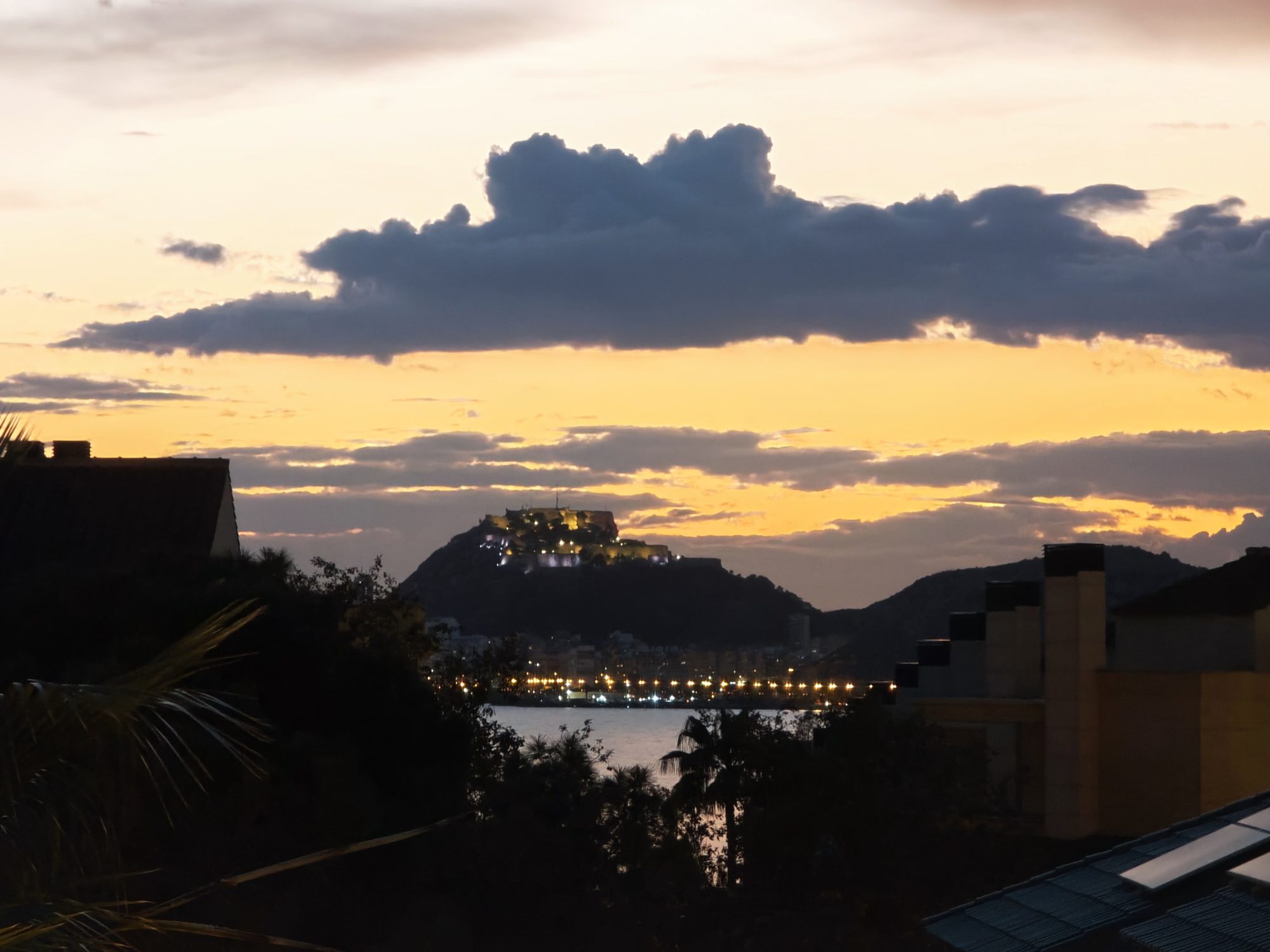 Castillo de Santa Bárbara iluminado al atardecer con cielo dorado y siluetas urbanas en Alicante.