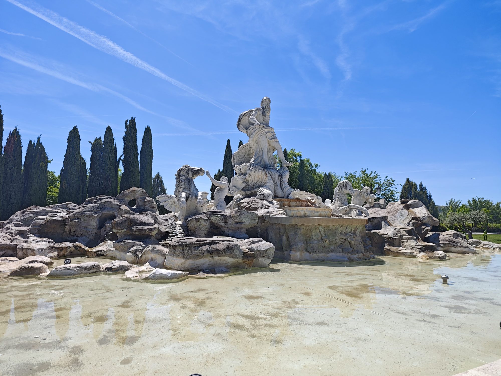 Fuente monumental con esculturas de Neptuno y caballos entre rocas bajo un cielo azul.
