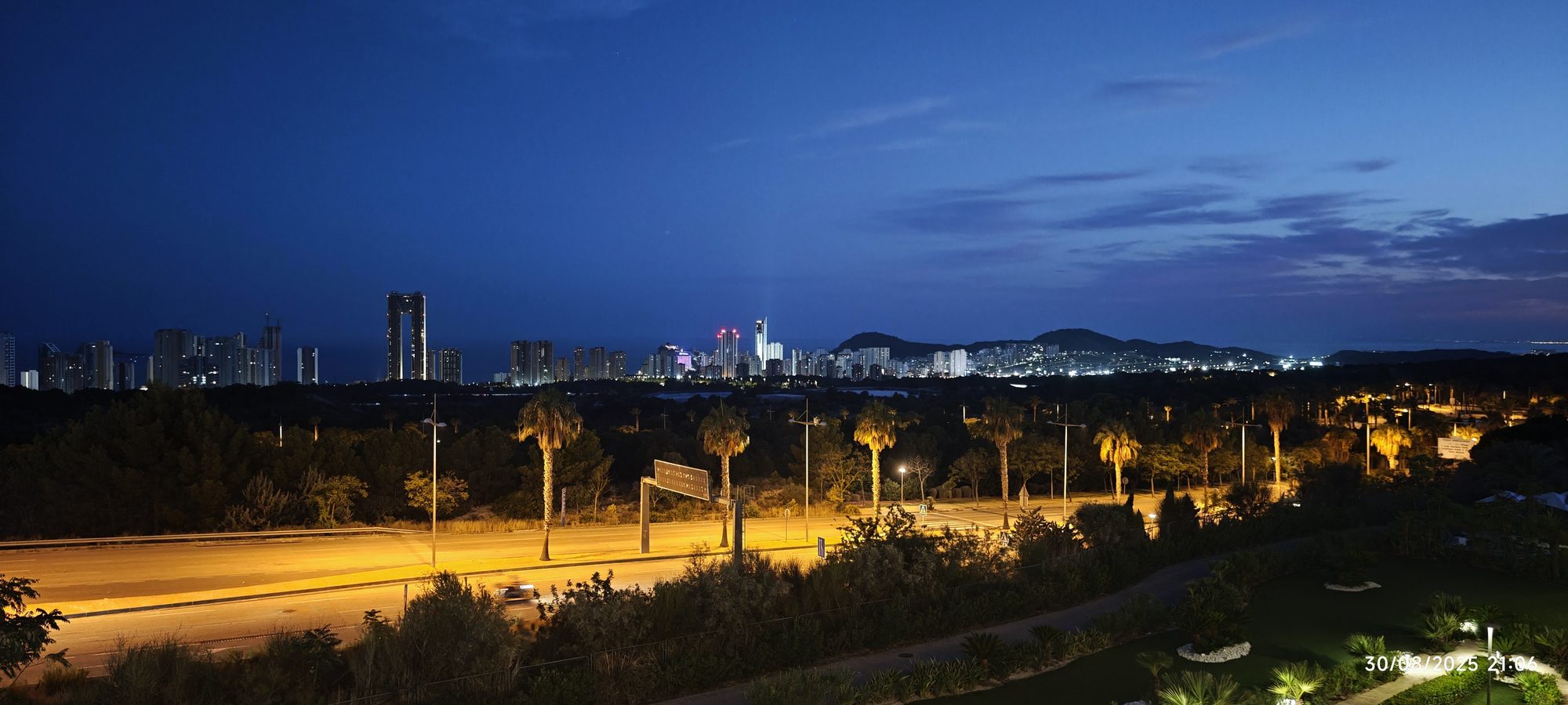 Skyline nocturno de Benidorm iluminado con rascacielos, palmeras y carretera bajo el cielo azul.