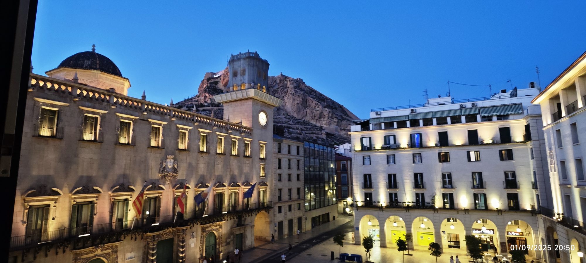Ayuntamiento de Alicante iluminado al anochecer con el Castillo de Santa Bárbara al fondo.