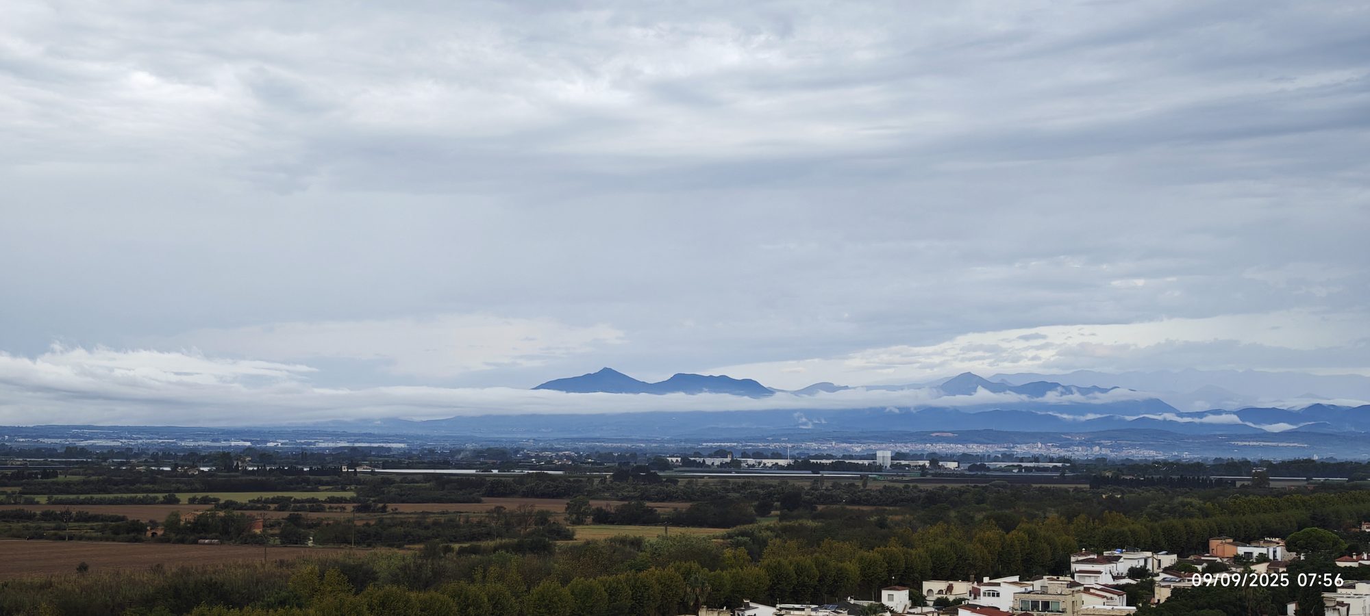Paisaje rural con campos de cultivo y montañas al fondo envueltas en nubes bajas.