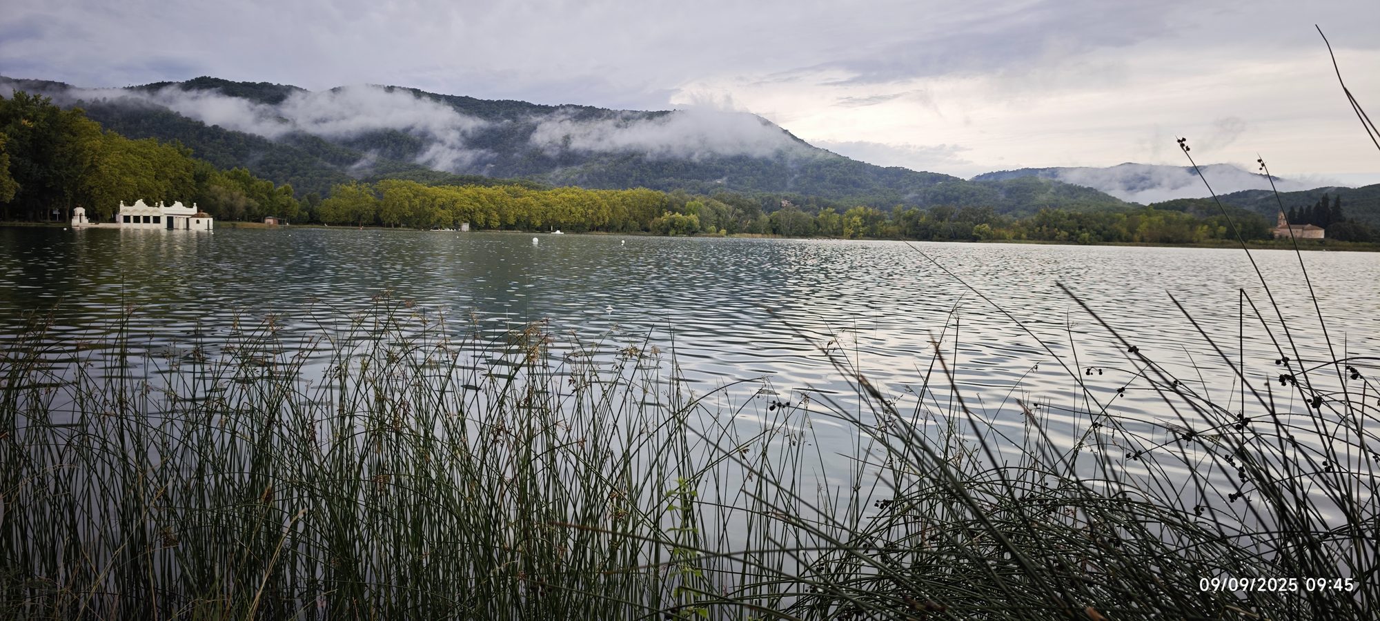 Lago de Banyoles rodeado de vegetación y montañas cubiertas por nubes bajas.