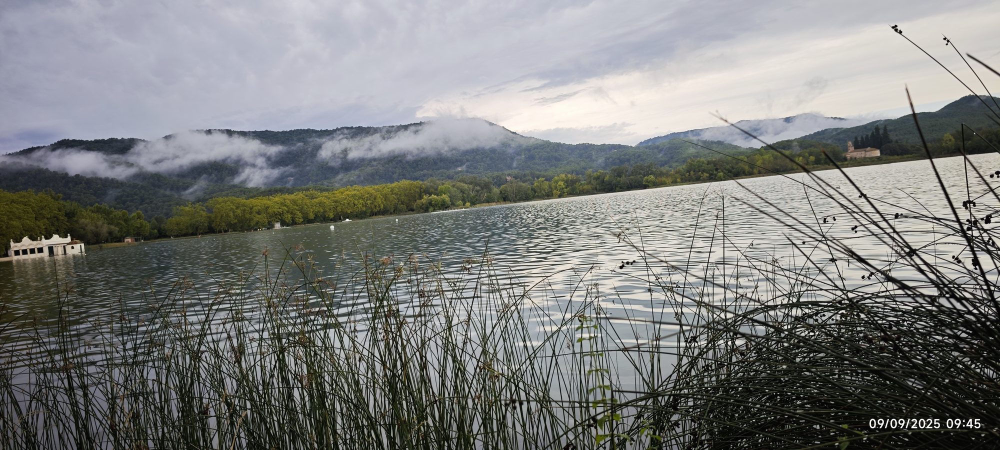 Lago de Banyoles entre cañaverales, montañas verdes y niebla baja en un día nublado.