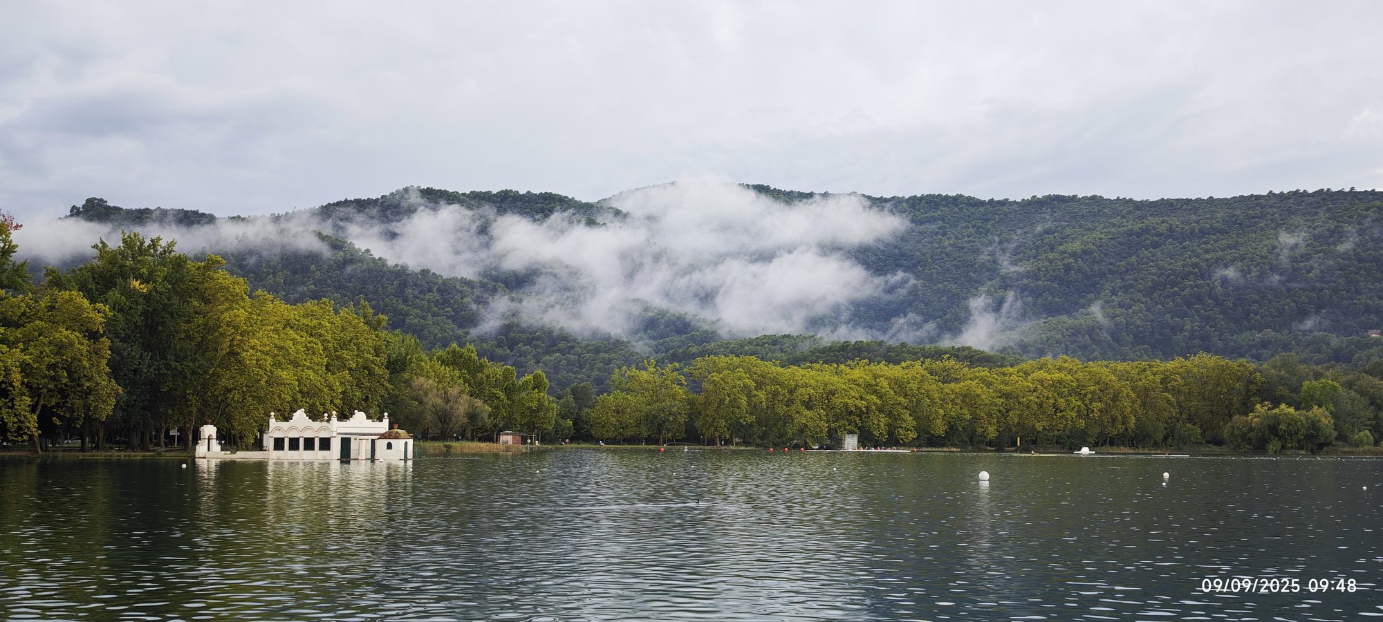 Lago de Banyoles con pesquera blanca, frondosos árboles y montañas cubiertas de niebla al fondo.