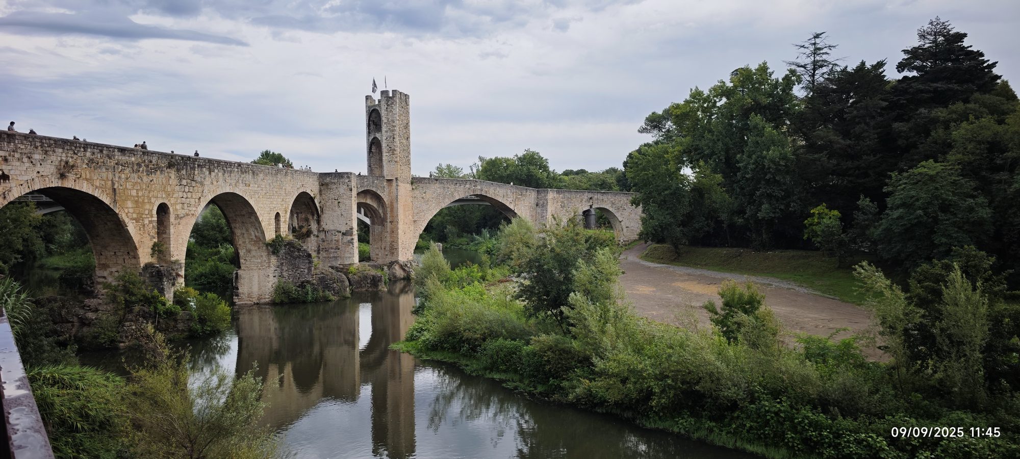 Puente medieval de piedra con torre fortificada sobre un río rodeado de vegetación verde.