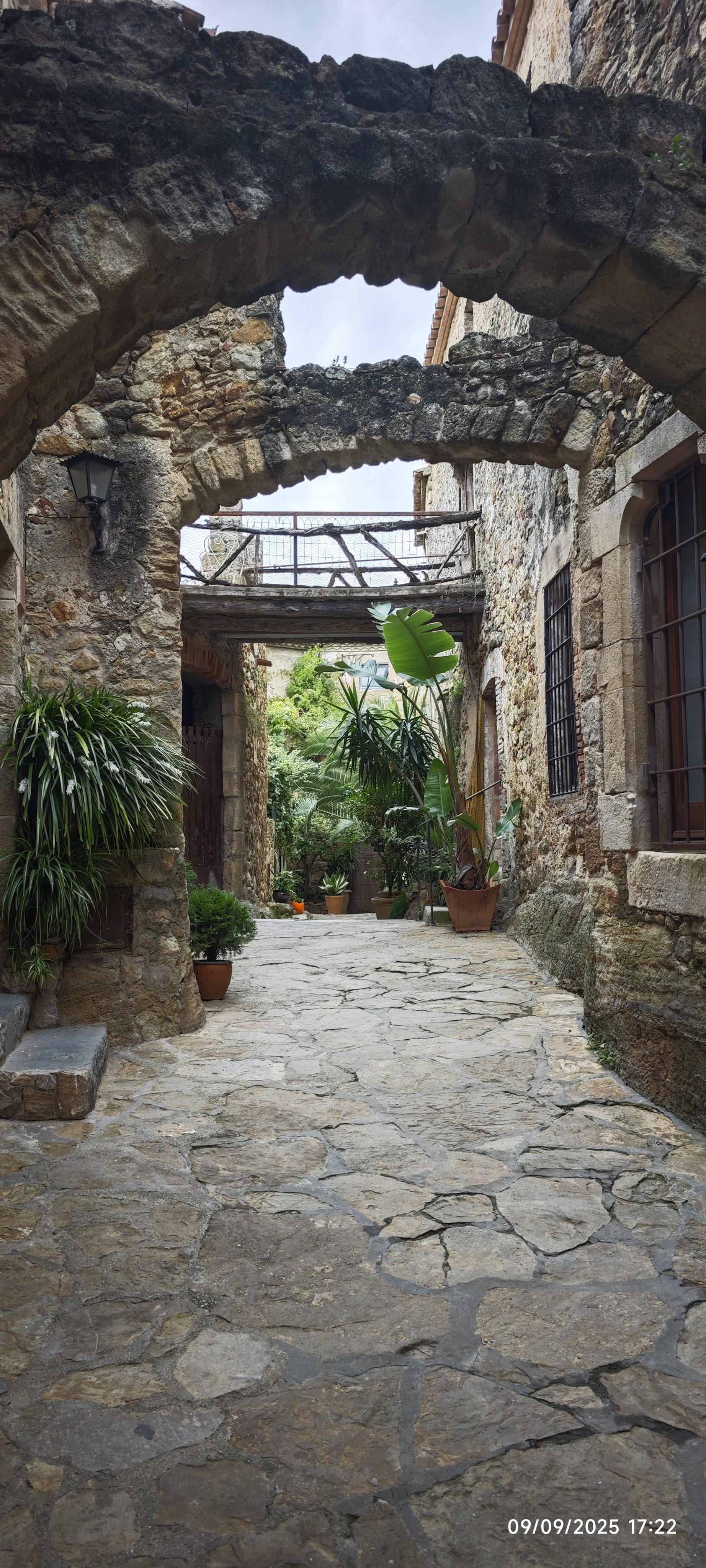 Calle empedrada medieval con arcos de piedra y plantas verdes en Pals, España.
