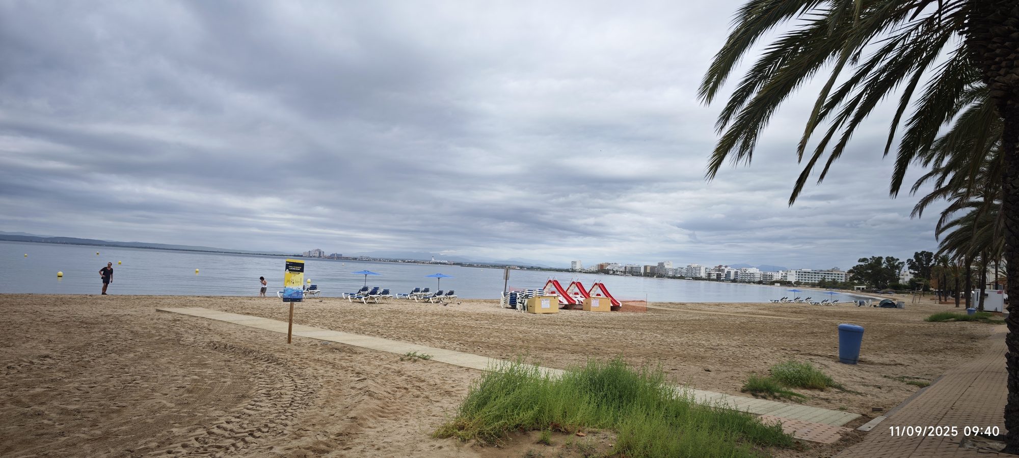 Playa de arena con sombrillas y palmeras bajo un cielo nublado junto al mar.