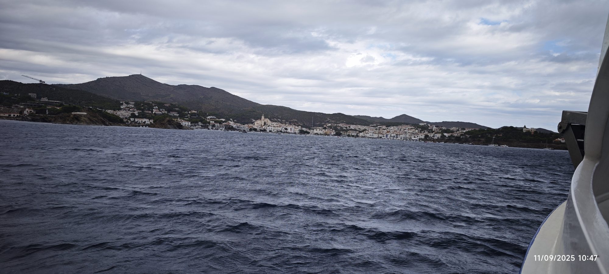 Vista costera de Cadaqués con casas blancas y montañas bajo cielo nublado desde el mar.