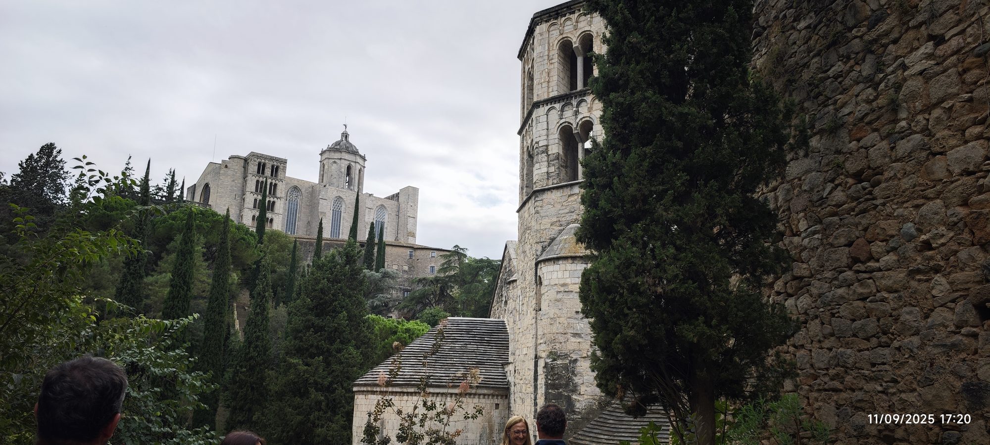 Torre de piedra románica y catedral de Gerona entre cipreses bajo un cielo nublado.