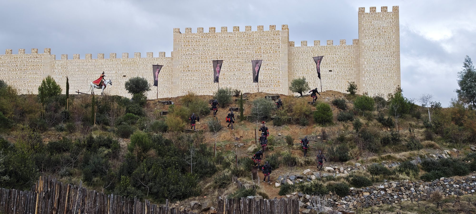 Muralla de piedra con estandartes y figuras de guerreros descendiendo por una colina.