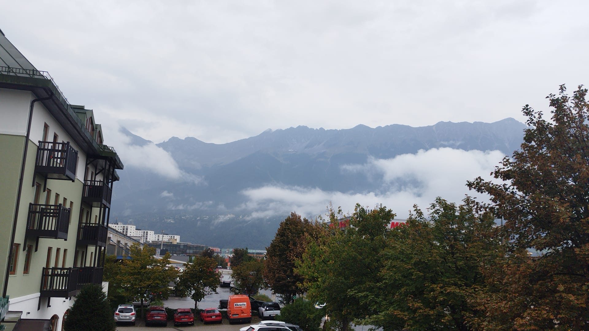 Montañas alpinas entre nubes bajas vistas desde una zona urbana con edificios y árboles.