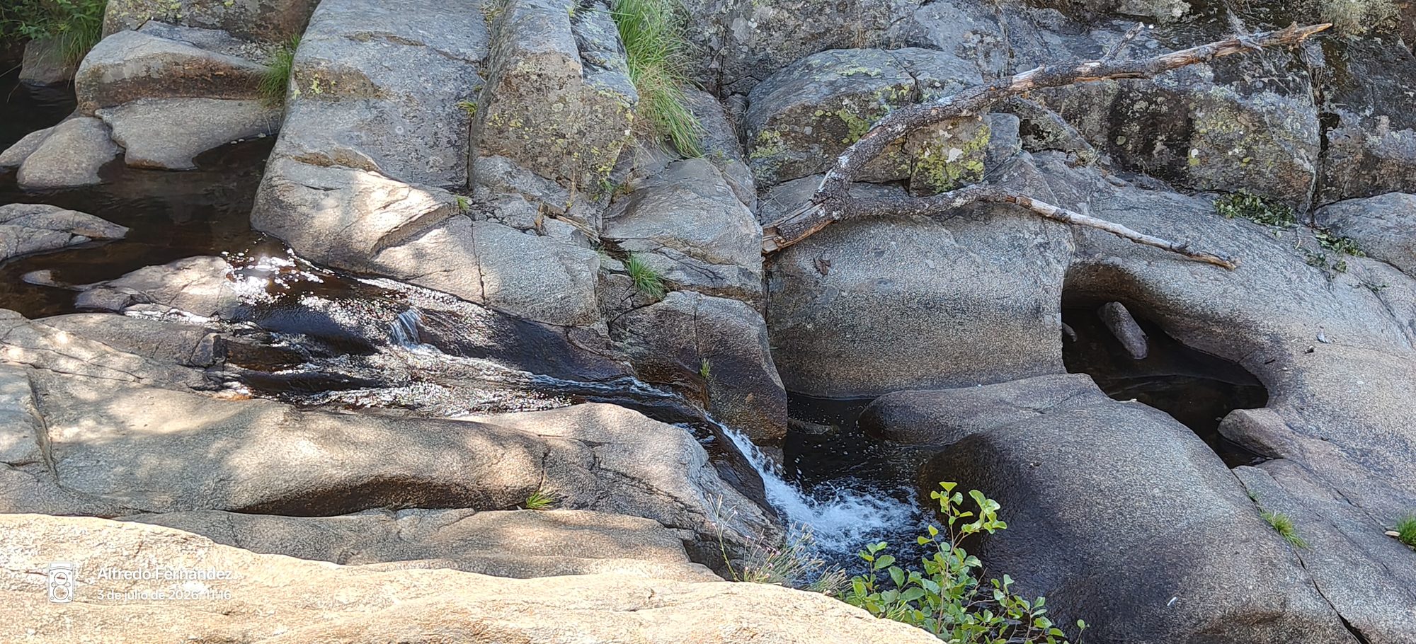 Arroyo con agua fluyendo entre grandes rocas de granito y vegetación silvestre.