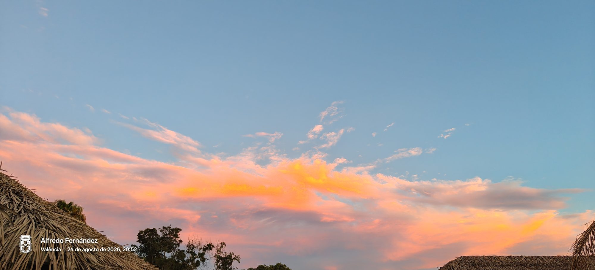 Nubes iluminadas con tonos naranjas y rosados al atardecer sobre techos de paja.