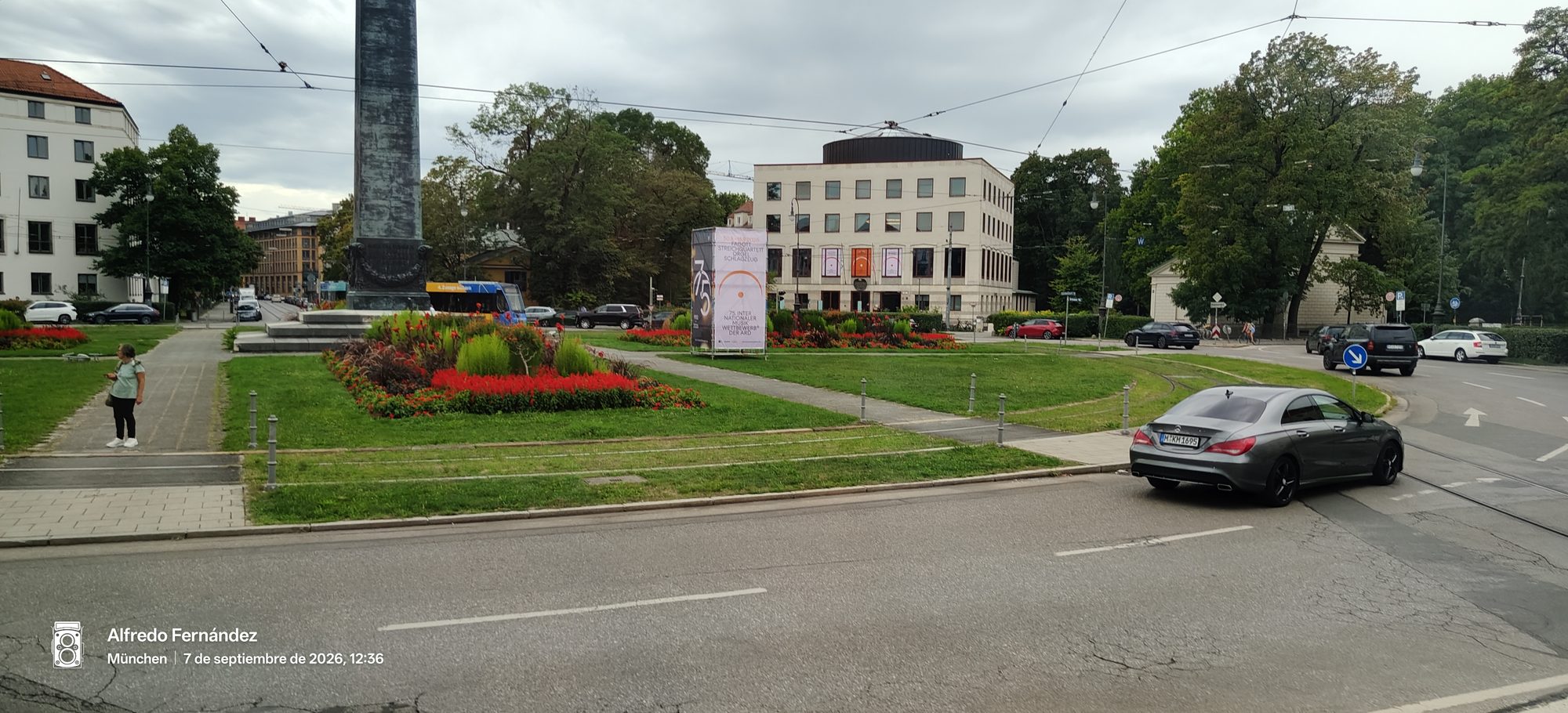 Plaza urbana en Múnich con jardines florales, base de obelisco y edificios al fondo.
