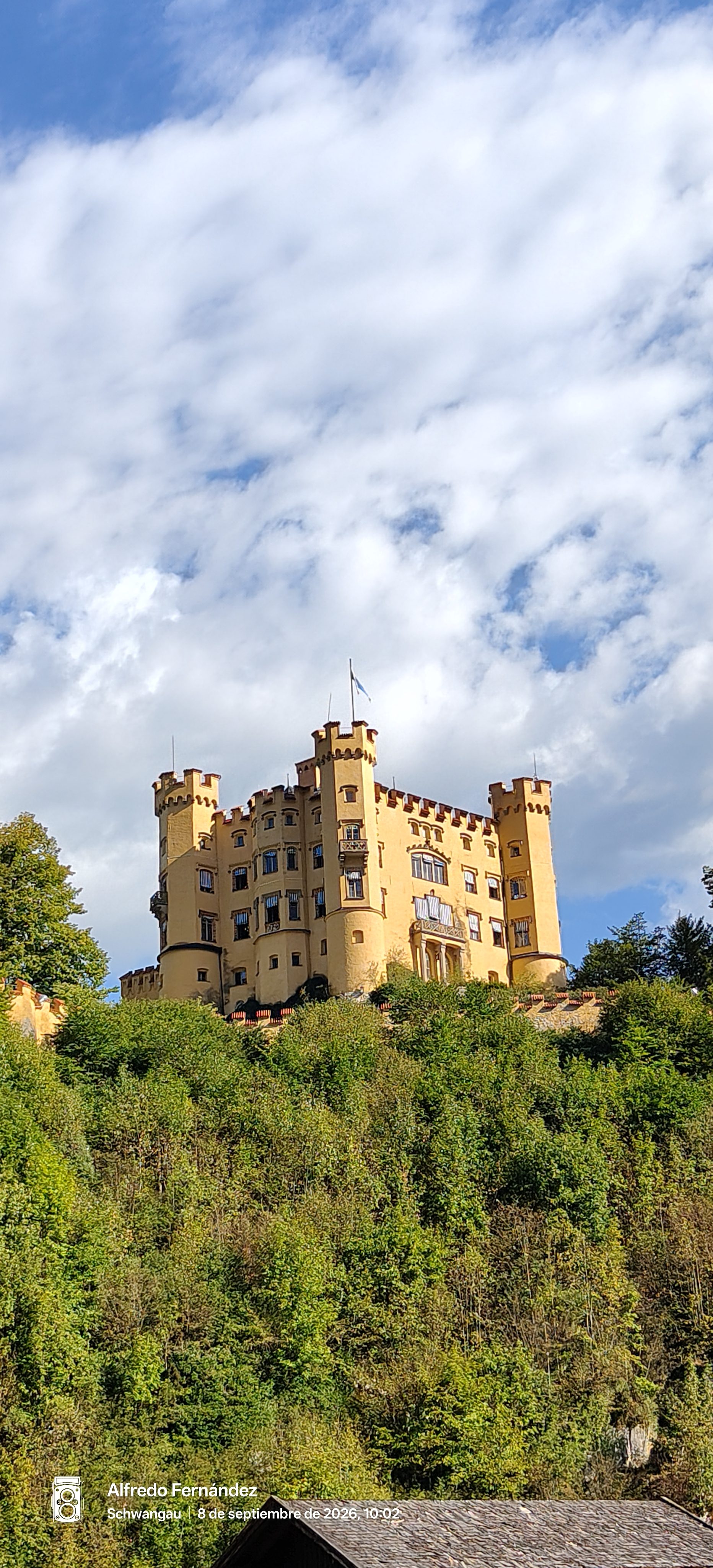 Castillo amarillo de Hohenschwangau entre colinas boscosas bajo un cielo con nubes.