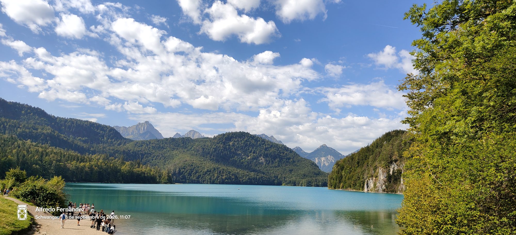 Lago de aguas turquesas rodeado de montañas boscosas y cielo con nubes en Schwangau.