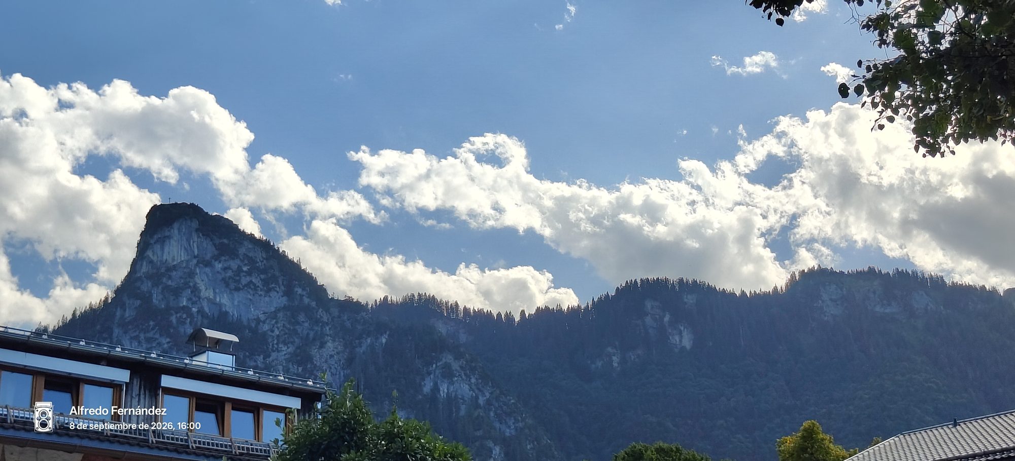 Montañas alpinas y cielo azul con nubes sobre el tejado de un edificio tradicional.
