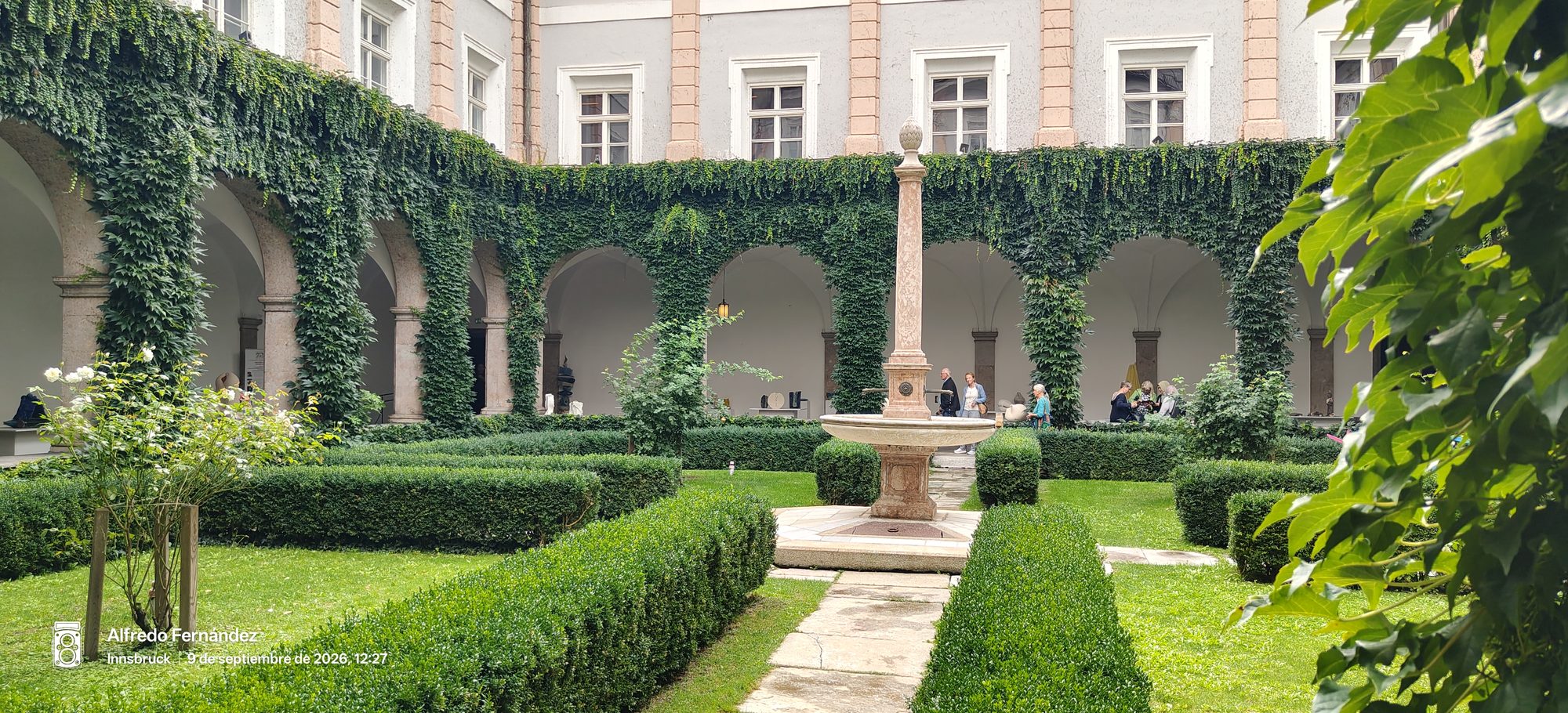 Patio ajardinado con fuente central de piedra y arcos cubiertos de frondosa hiedra.