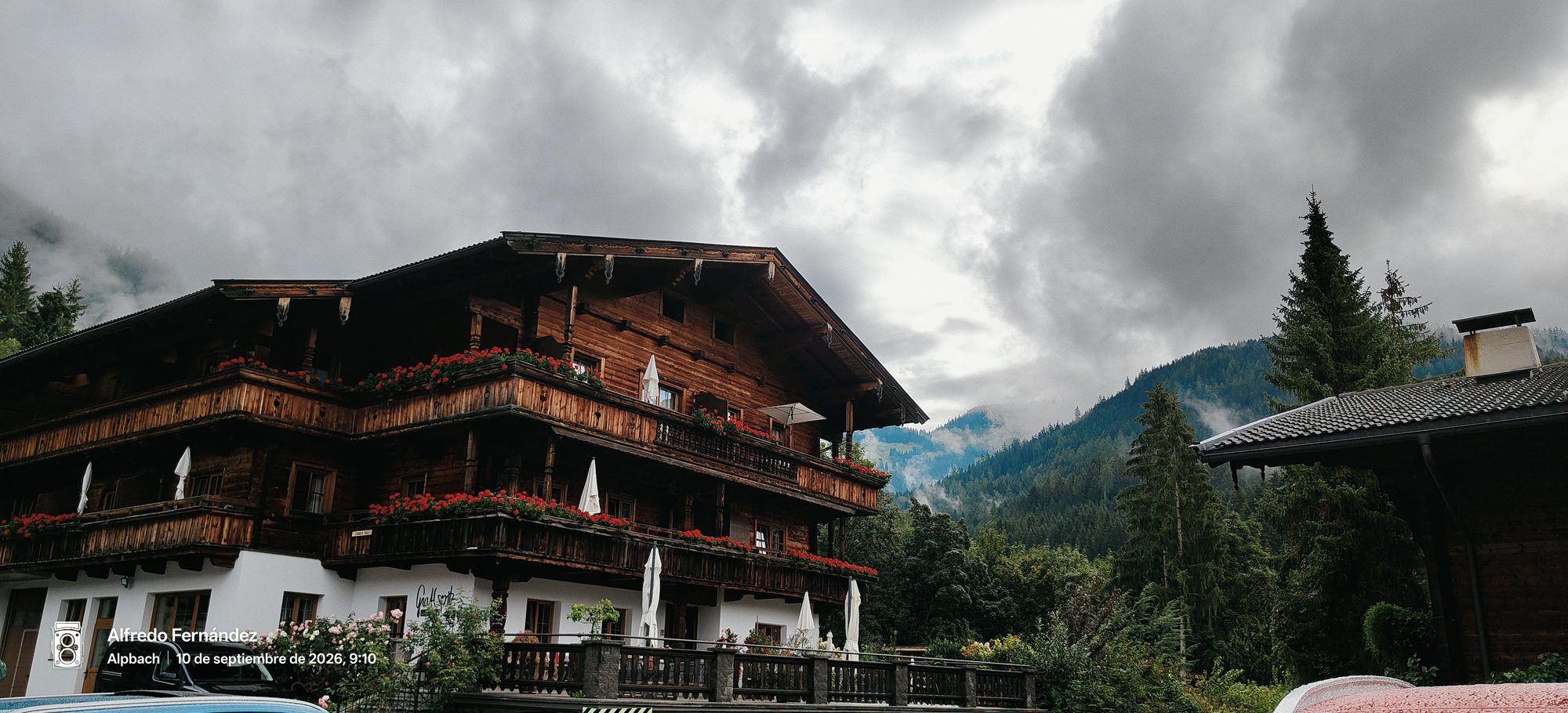 Chalet tradicional de madera con balcones florecidos frente a montañas alpinas con niebla.