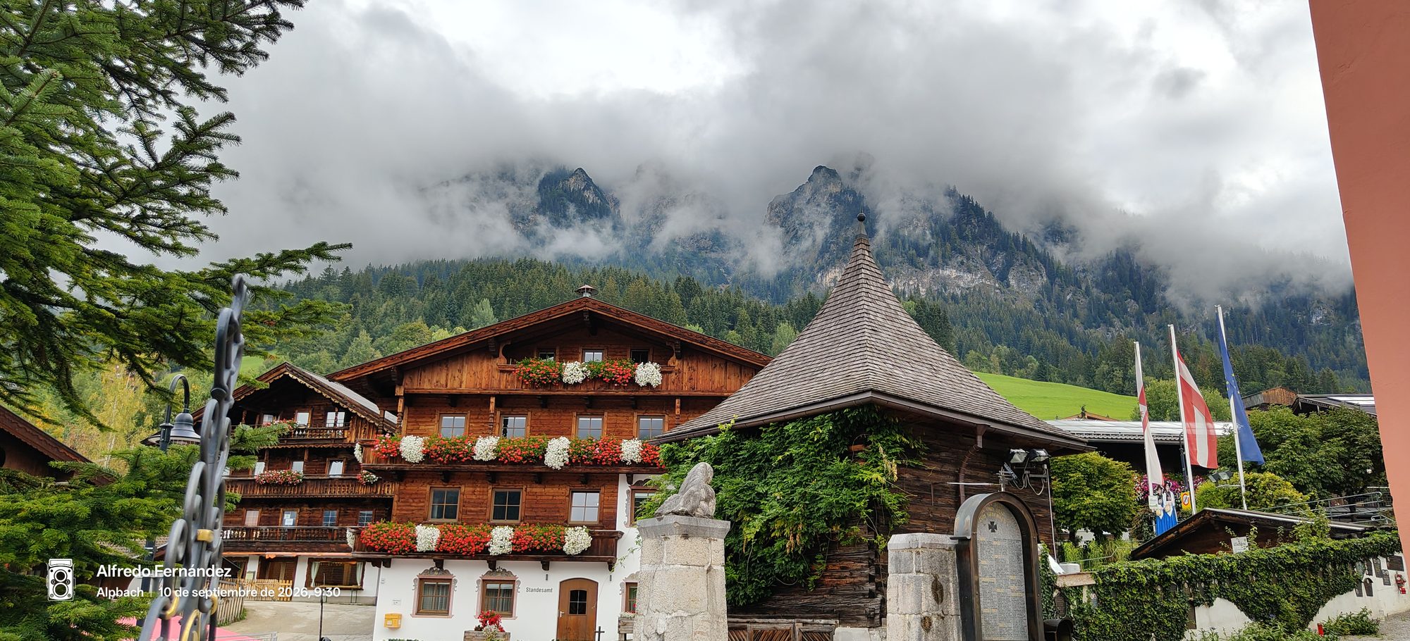 Chalets tradicionales de madera con balcones floridos frente a montañas alpinas entre niebla.