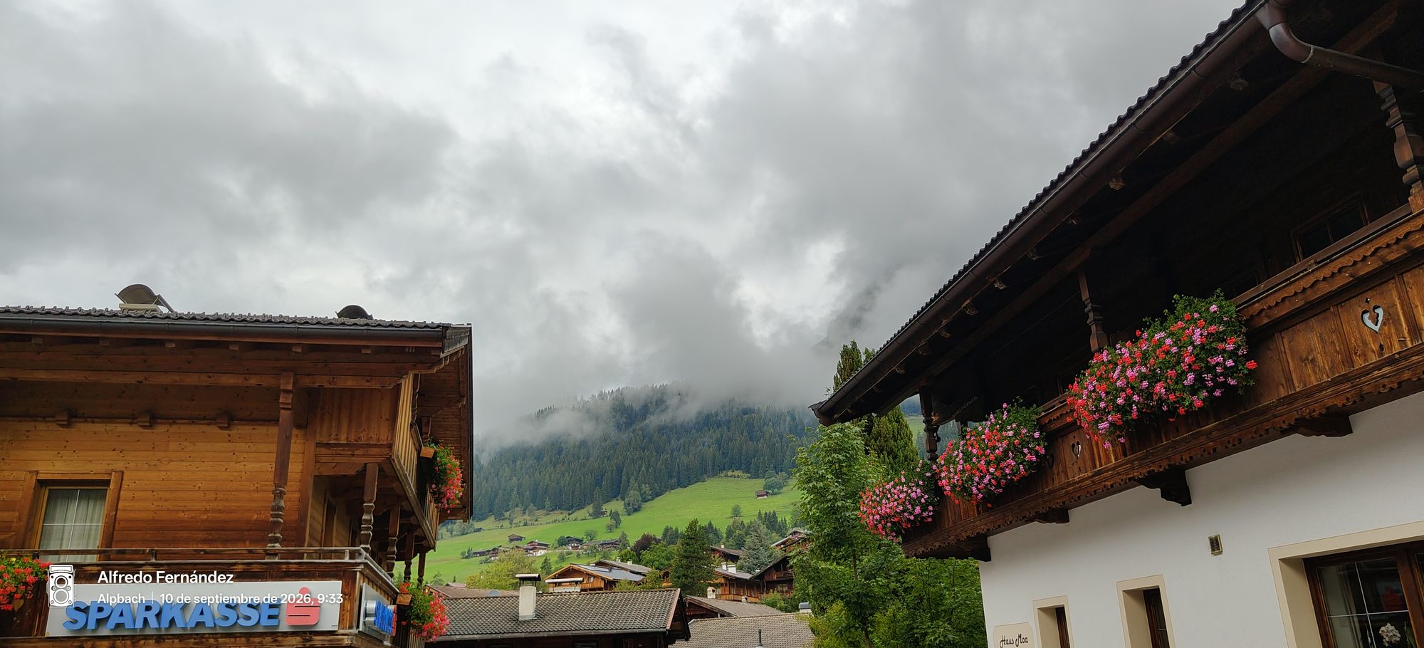 Casas alpinas de madera con balcones floridos y montañas verdes cubiertas de niebla.