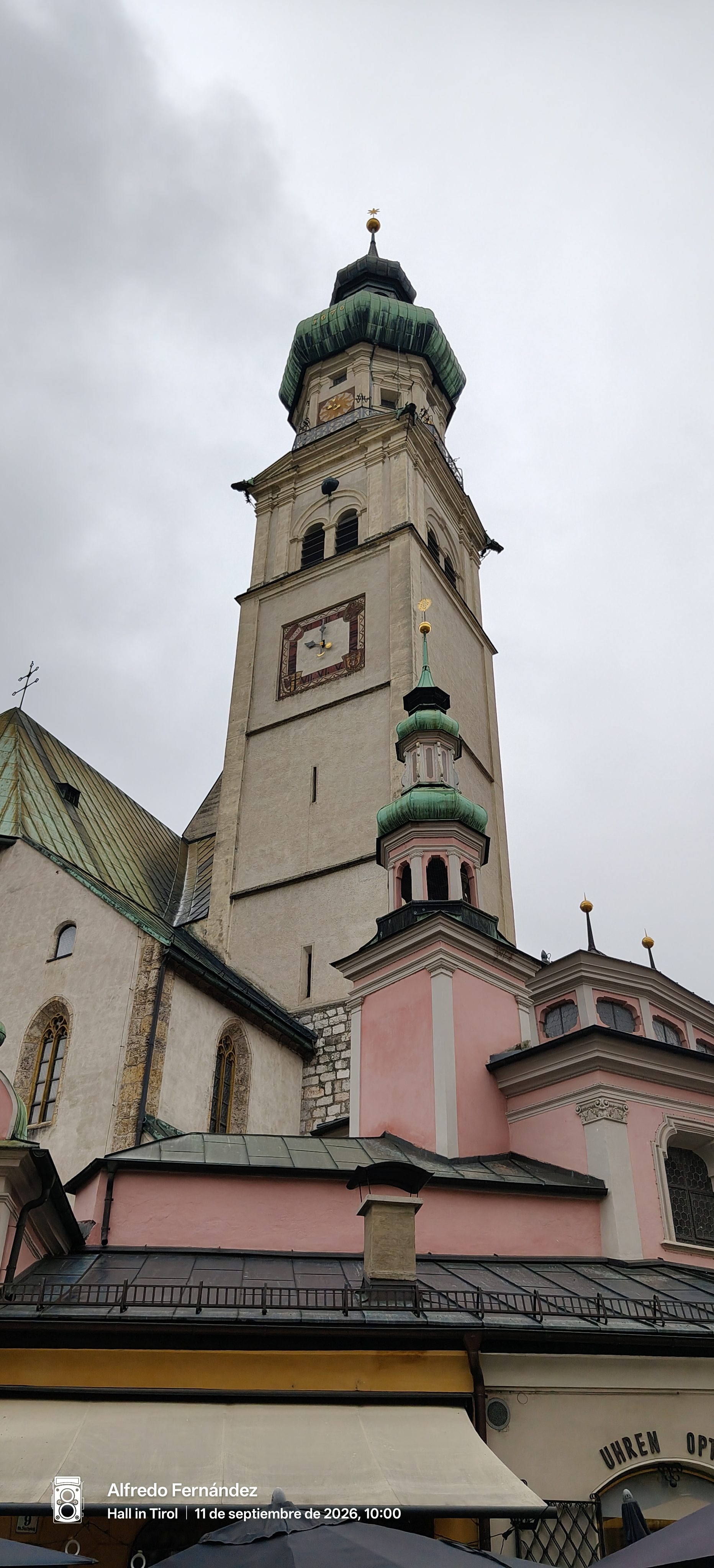Torre campanario con cúpula de cobre y capilla barroca rosa bajo cielo nublado.