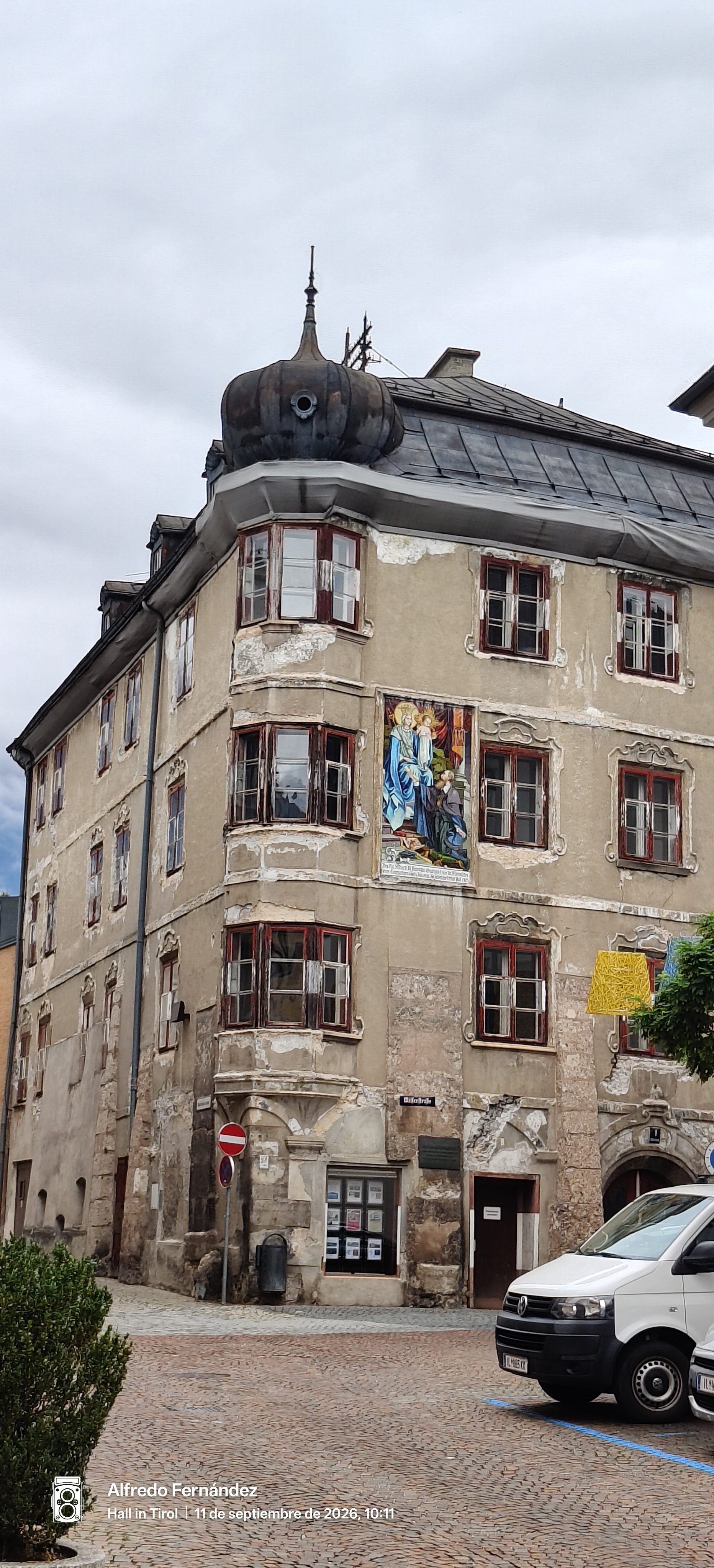 Edificio histórico con miradores de madera y fresco religioso en una plaza adoquinada.