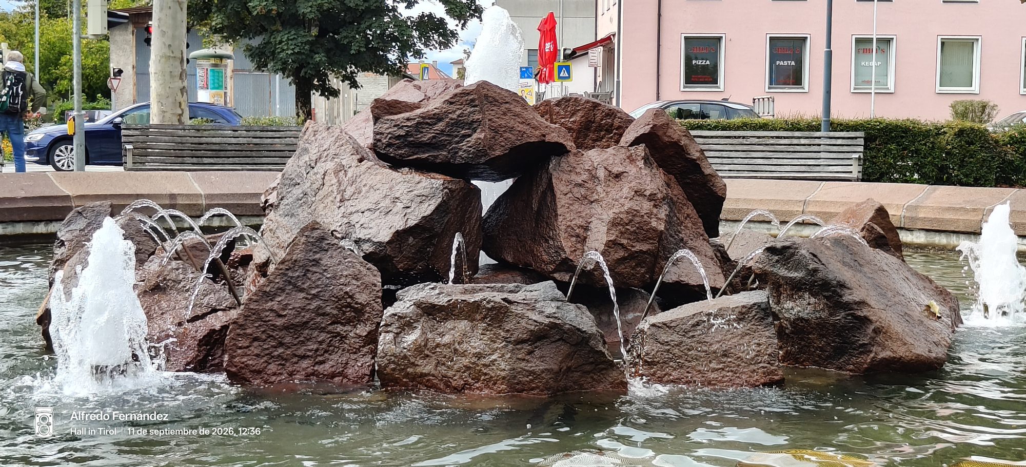 Fuente urbana con grandes rocas rojizas y chorros de agua en una plaza.