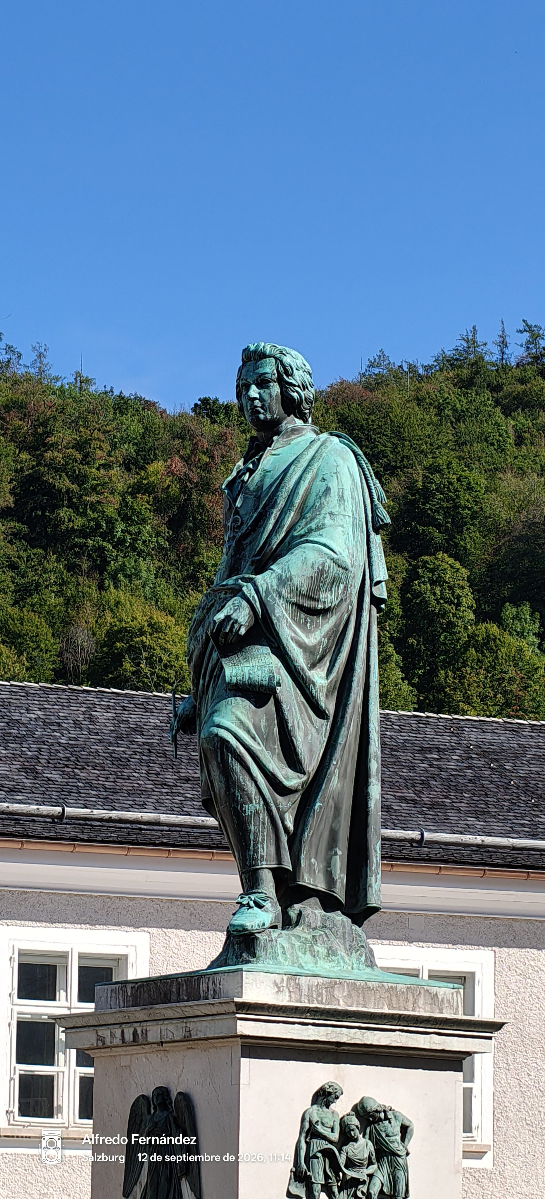 Estatua de bronce de Mozart sobre pedestal en Salzburgo con colina verde al fondo.