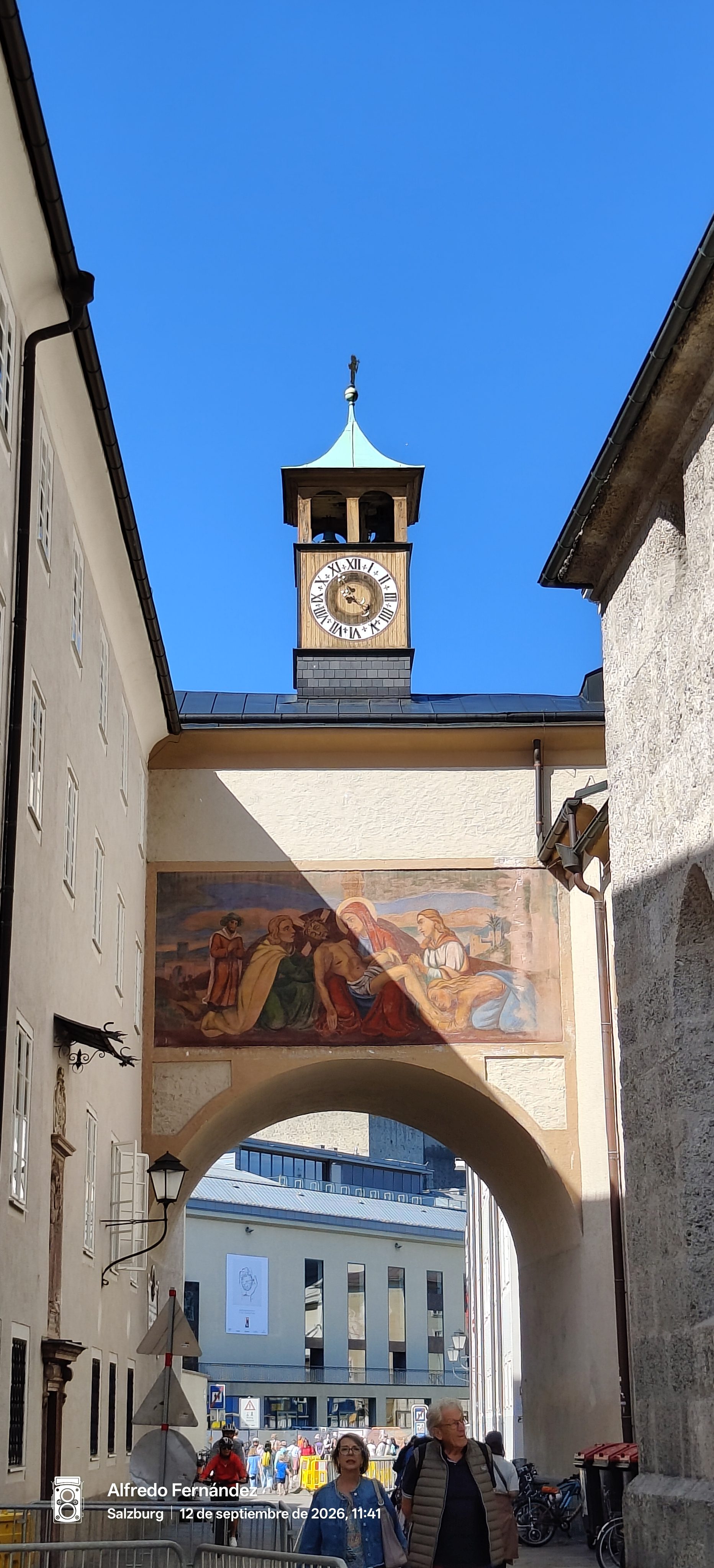 Arco histórico con fresco religioso y torre del reloj bajo un cielo despejado.