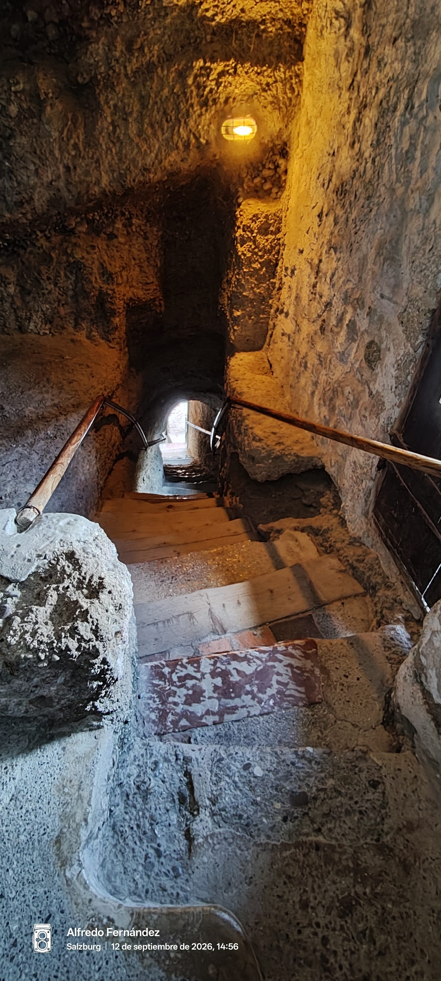 Escalera estrecha de piedra en un túnel subterráneo iluminado con tenue luz cálida.