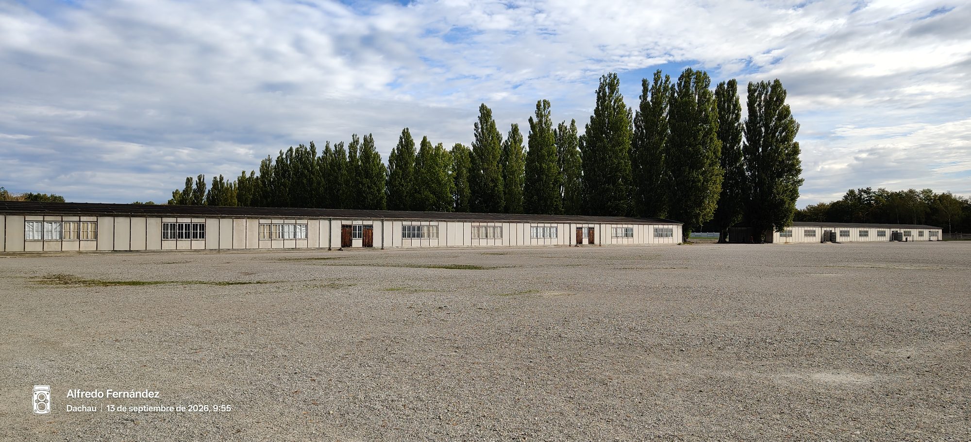 Barracones reconstruidos frente a una amplia explanada de grava y álamos en Dachau.