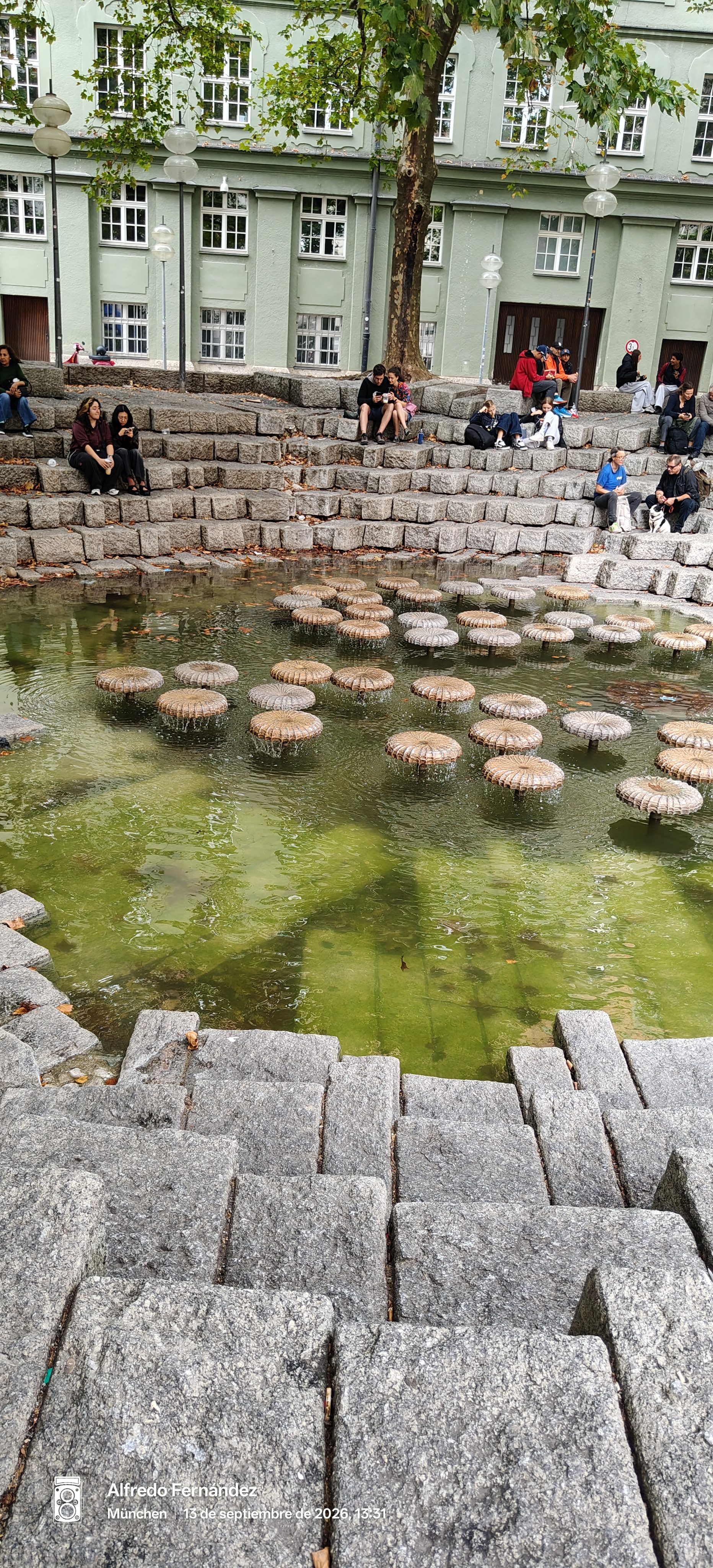 Estanque urbano con fuentes circulares en forma de hongo y gradas de piedra.