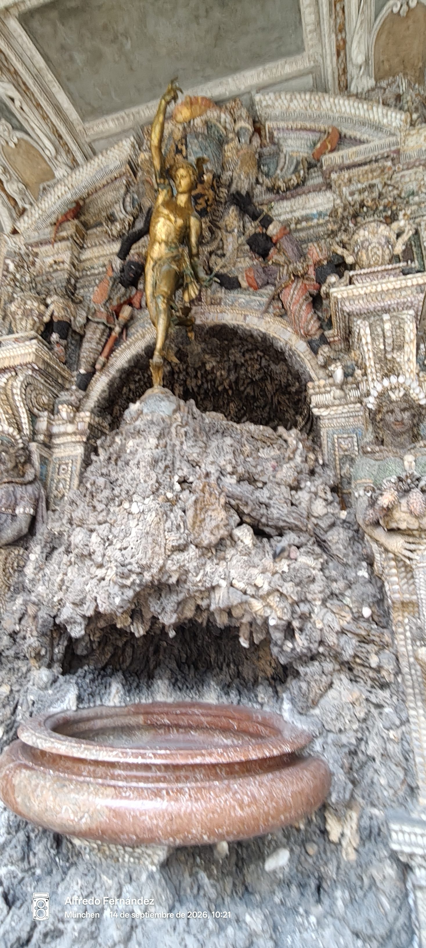 Gruta decorada con conchas marinas, fuente de piedra y estatua dorada en su cima.