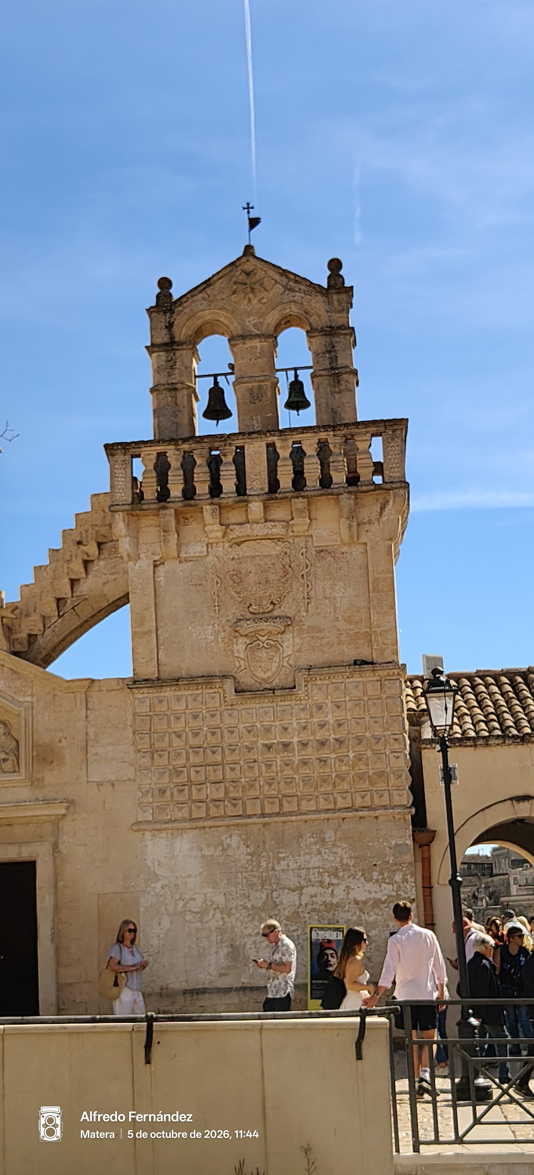 Espadaña de piedra con campanas y relieves tallados bajo un cielo azul despejado.