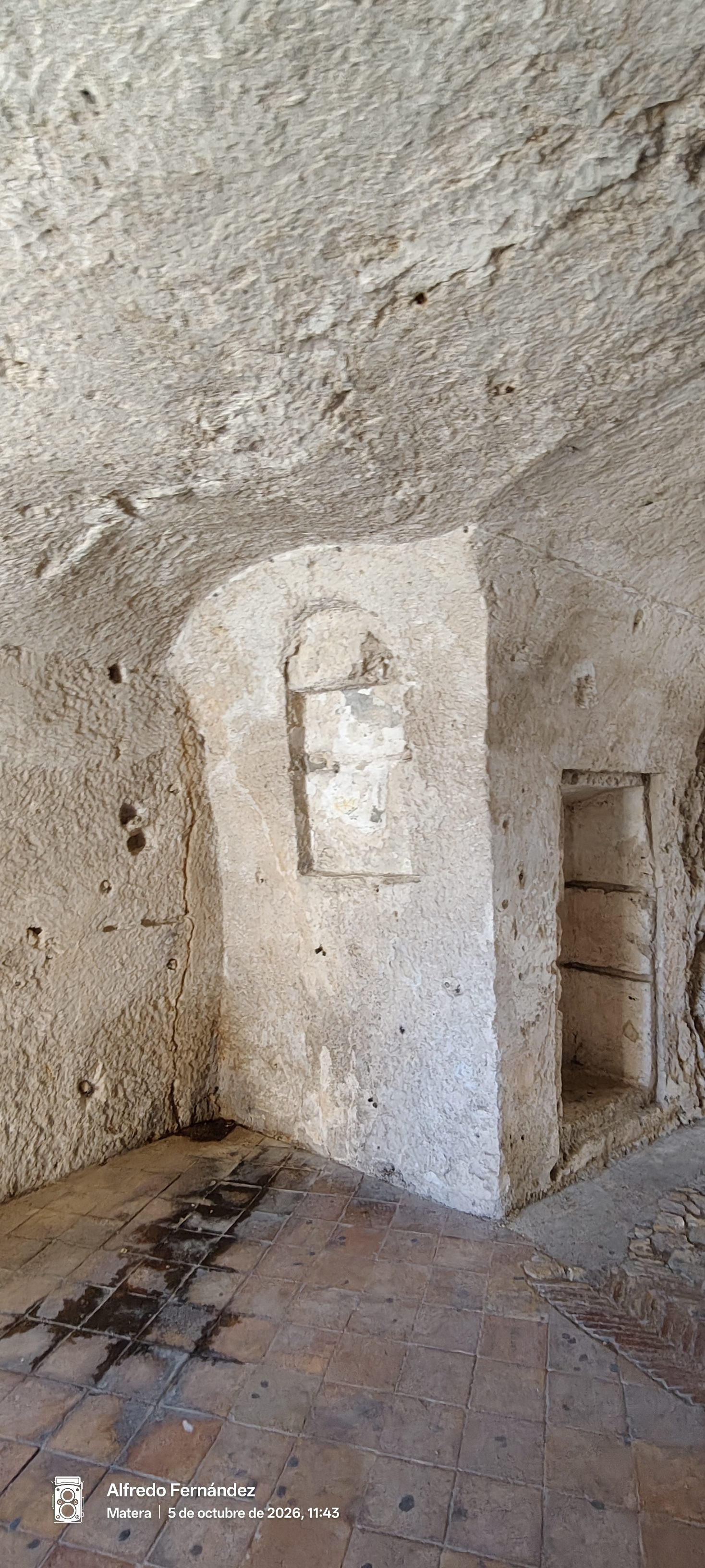 Interior de una estancia rupestre excavada en roca con nichos antiguos y suelo de baldosas.