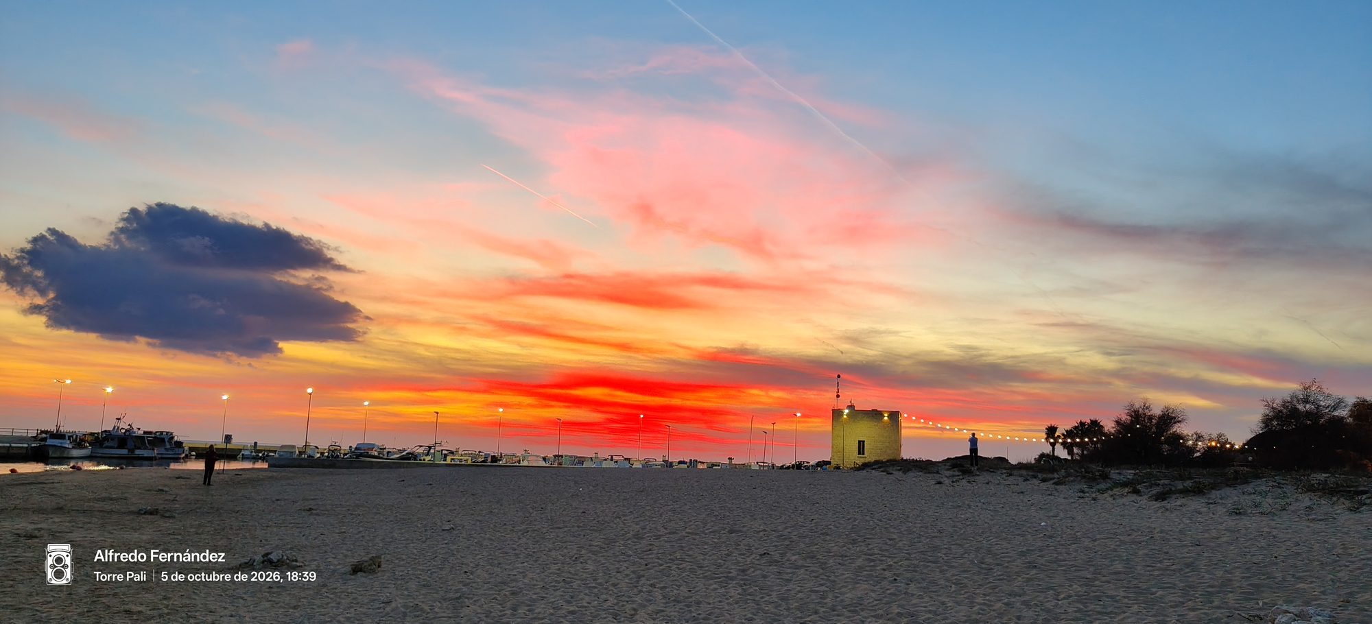 Atardecer rojizo sobre la playa de arena y el pequeño puerto con barcos.
