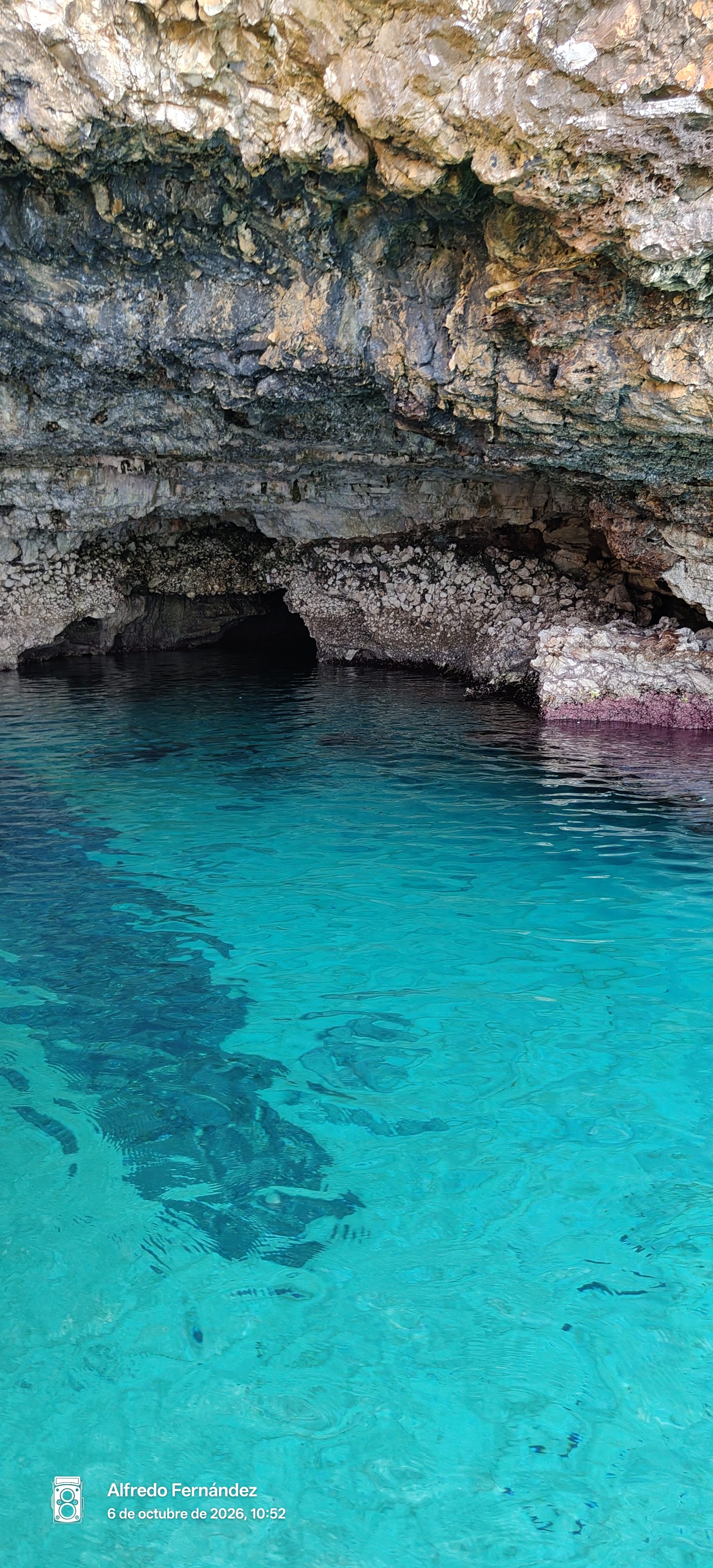 Aguas turquesas transparentes frente a la entrada rocosa de una cueva marina.