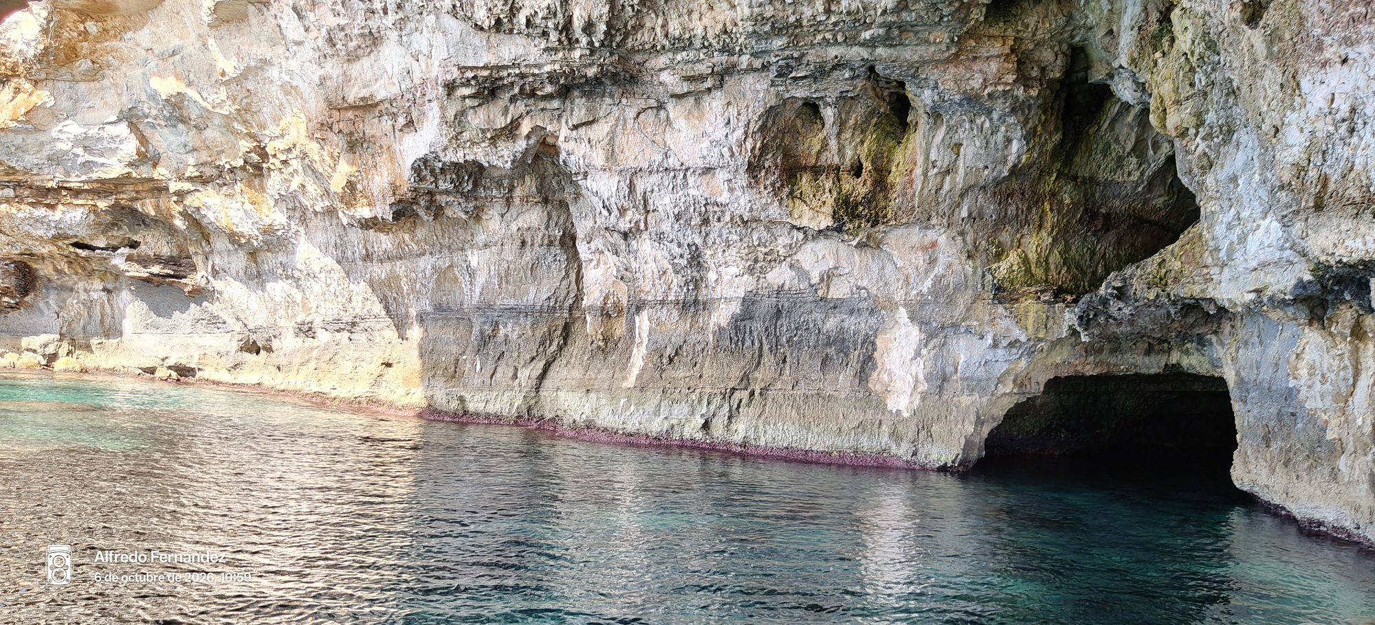 Acantilado de roca caliza y entrada a una cueva marina sobre aguas cristalinas.