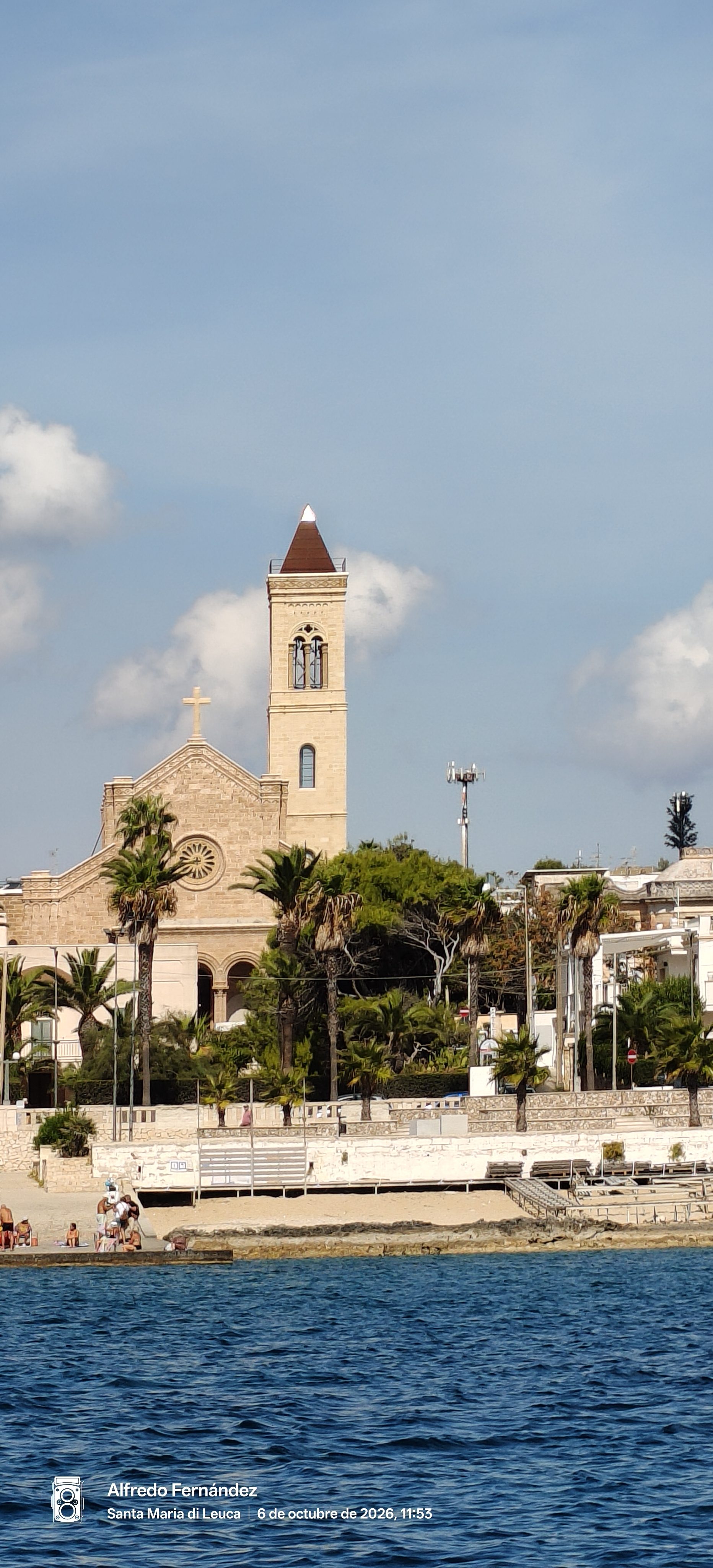 Iglesia de piedra con alto campanario y palmeras frente al mar azul.