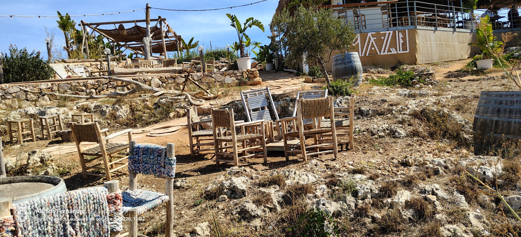 Sillas rústicas de madera en una terraza costera y rocosa bajo el sol mediterráneo.