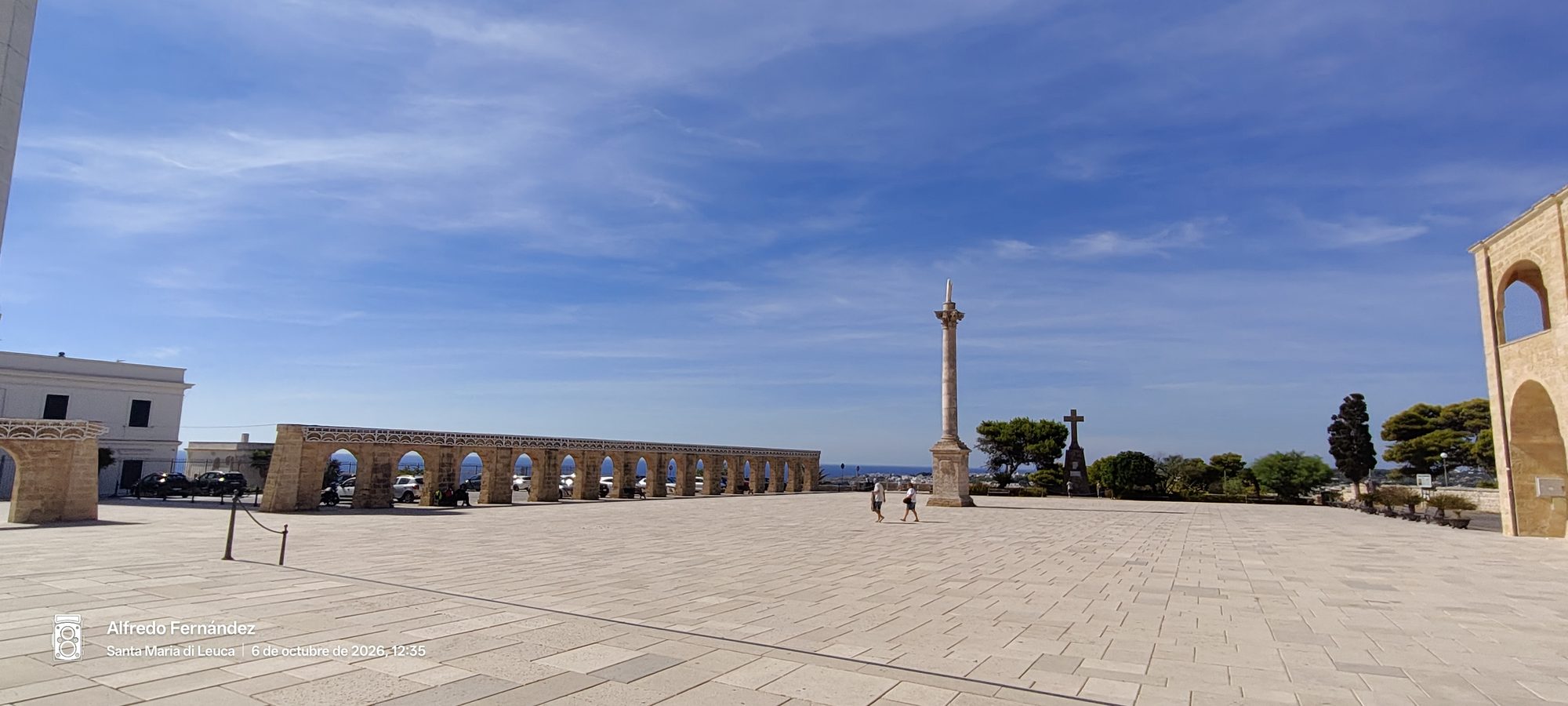 Amplia plaza empedrada con arcadas y columna monumental bajo un cielo azul despejado.