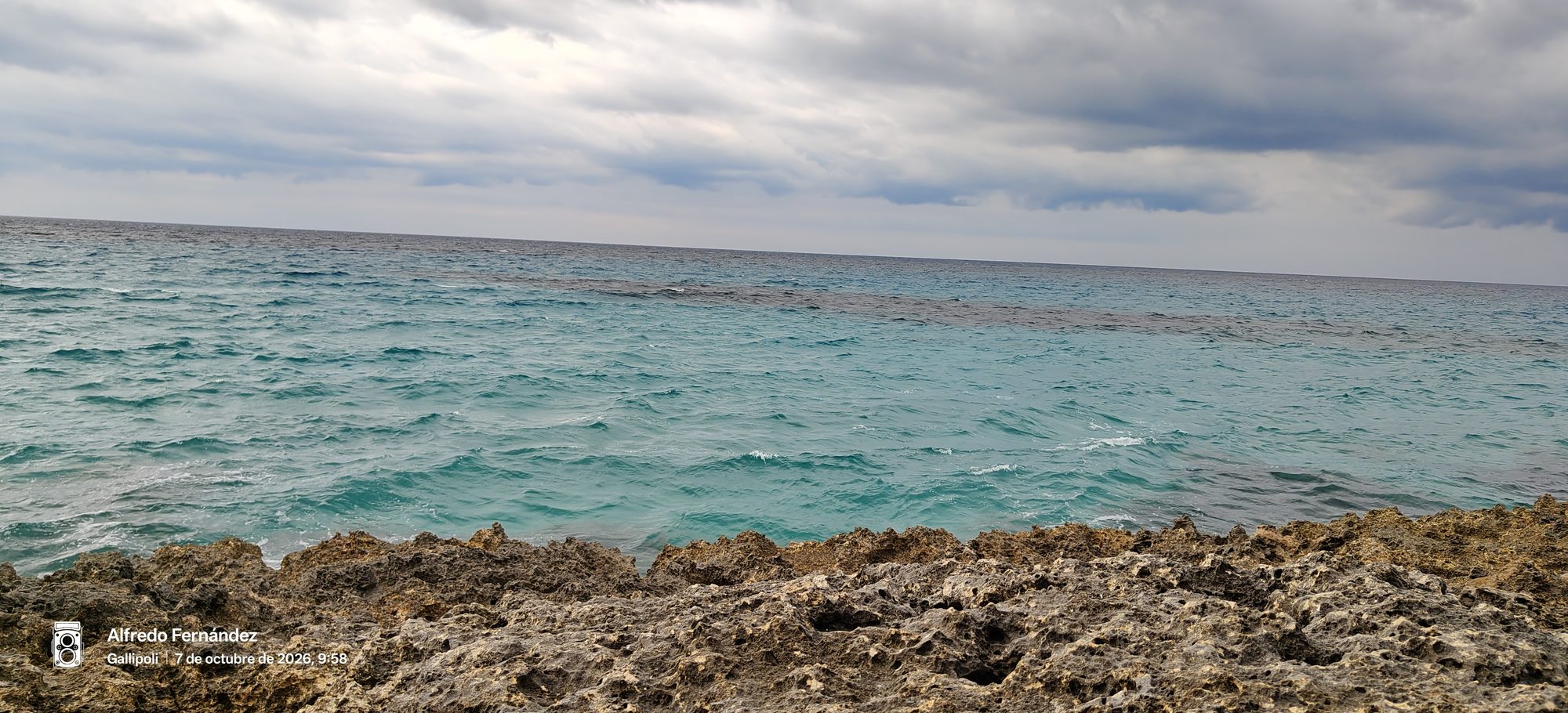 Mar turquesa ondulado visto desde una costa rocosa bajo un cielo nublado y gris.
